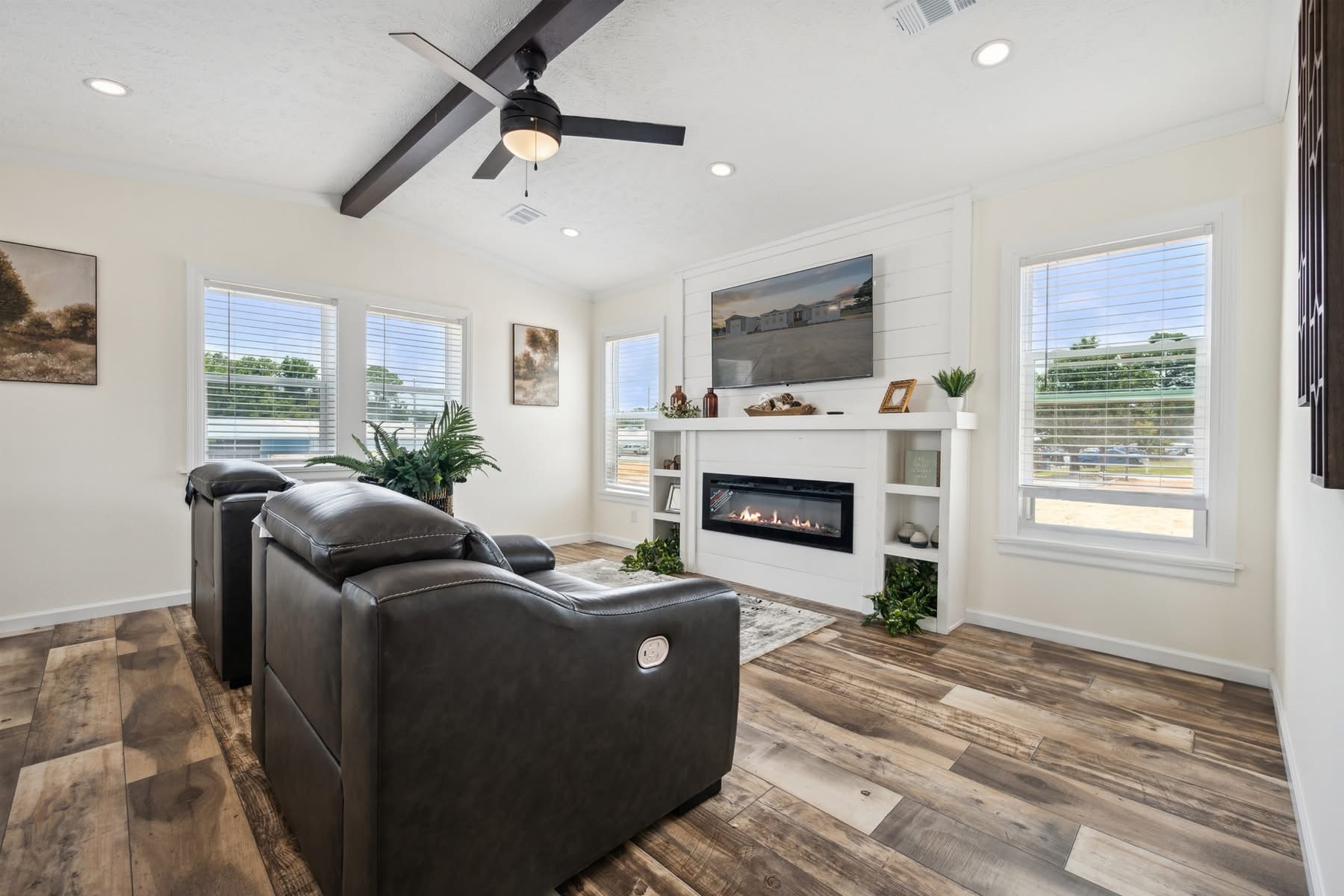 Cozy living room with dark leather recliners facing a modern fireplace beneath landscape art. Large windows brighten the room with natural light.