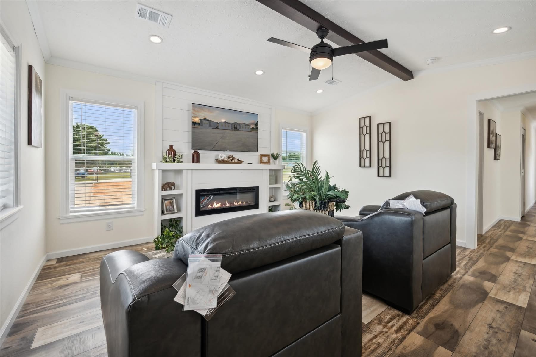 Modern living room with two gray armchairs facing a fireplace and TV. The room features large windows, wooden floors, and a ceiling fan, creating a cozy ambiance.