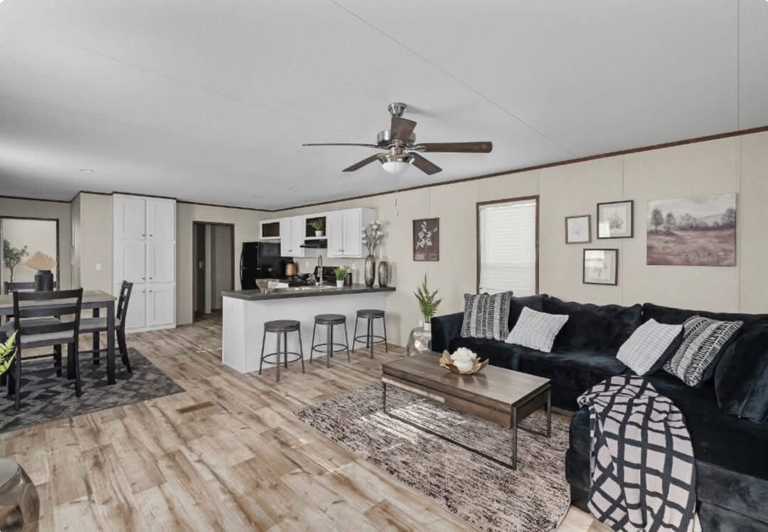 A modern living area with wooden flooring, featuring a dark sofa with cushions, a coffee table, and a ceiling fan. A kitchen with a breakfast bar and stools is visible in the background, along with a dining area with a table and chairs. The decor is neutral with framed artwork and plants.