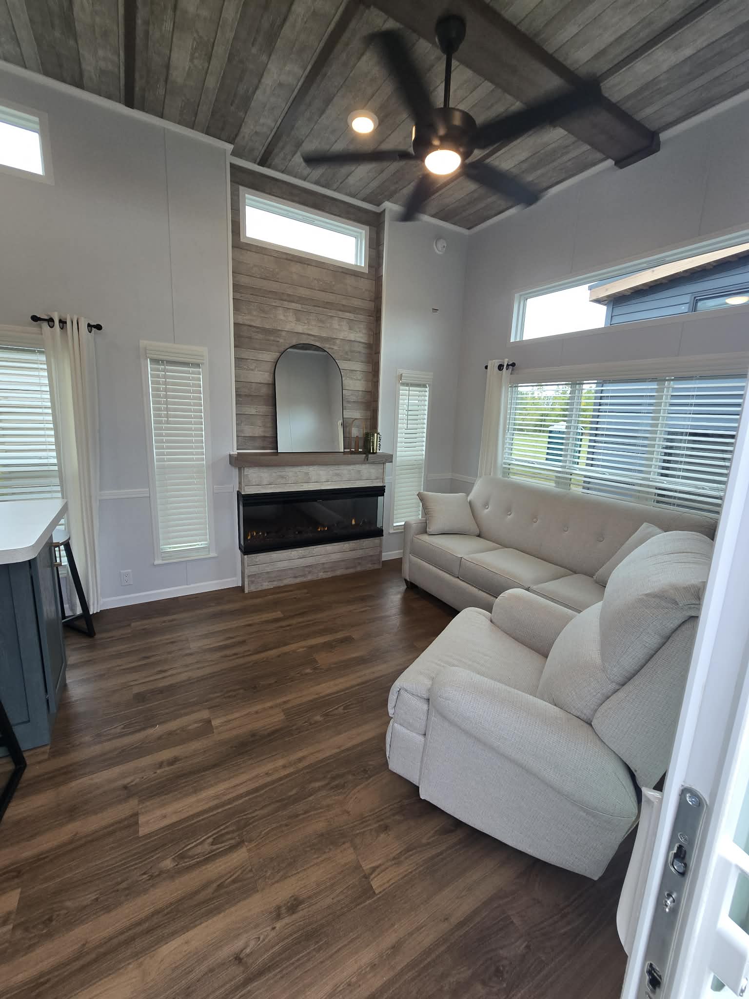 Cozy living room with wood flooring, beige sectional sofa, and armchair. Features a stone fireplace, large mirror, and ceiling fan, conveying warmth.
