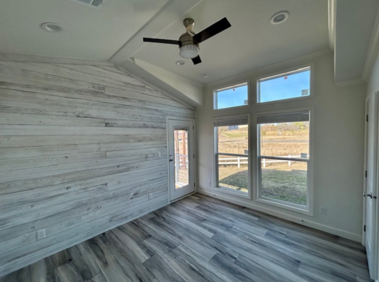 A small, empty room with white walls and light wood flooring. A lone window with closed blinds lets in soft light, giving a calm and minimalist feel.
