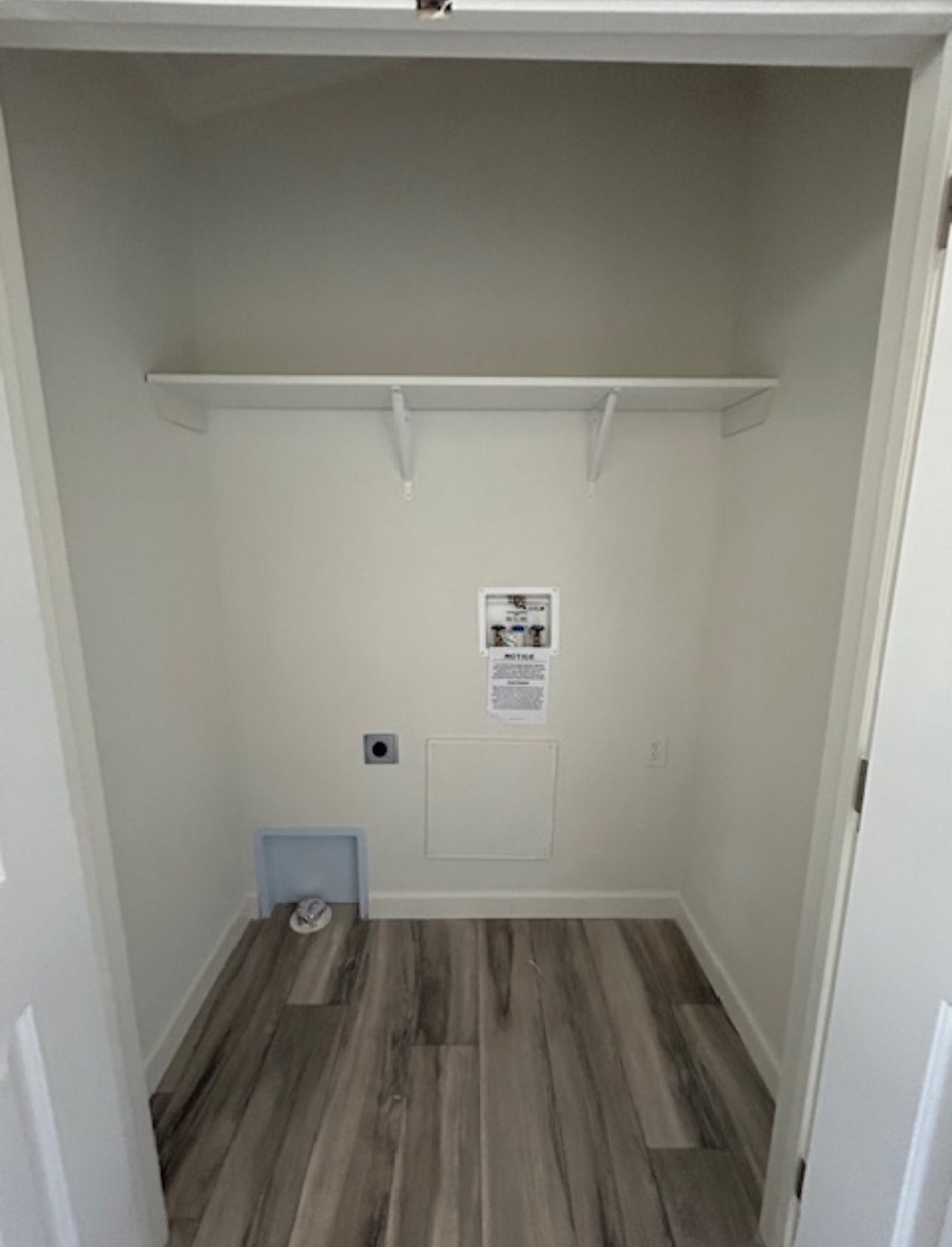 Empty laundry closet with light wood flooring, white walls, and shelf above hookups for washer and dryer. Notices are posted on the wall.