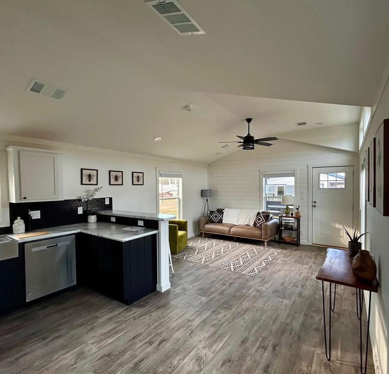 Modern living space with open layout. Features a sleek kitchen with dark cabinets, cozy beige sofa, patterned rug, green armchair, and bright windows.