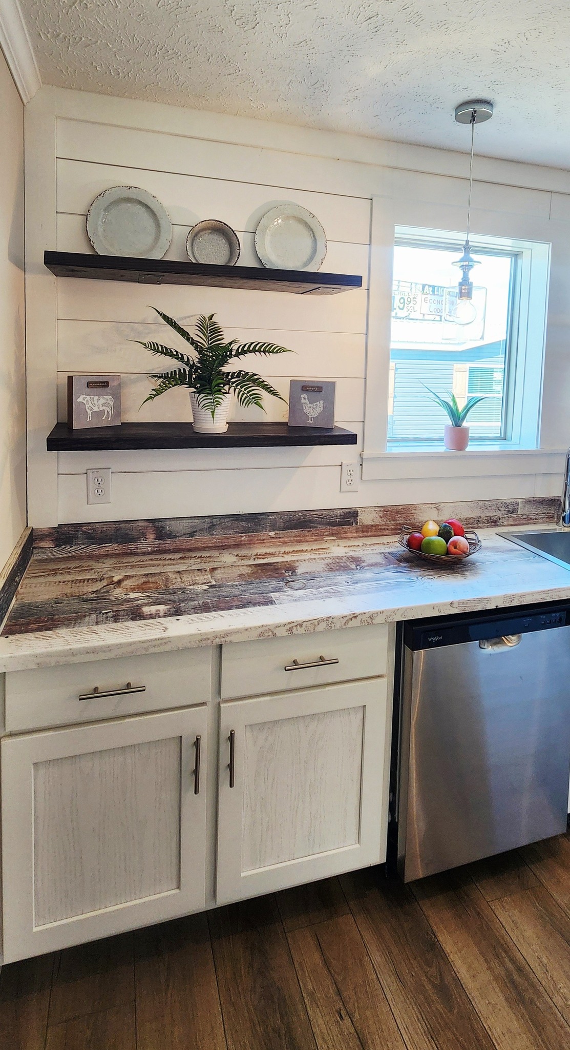 Simple kitchen with wooden shelves, farmhouse decor, and a rustic countertop. Fresh fruit in a bowl, a potted plant by the window adds warmth.