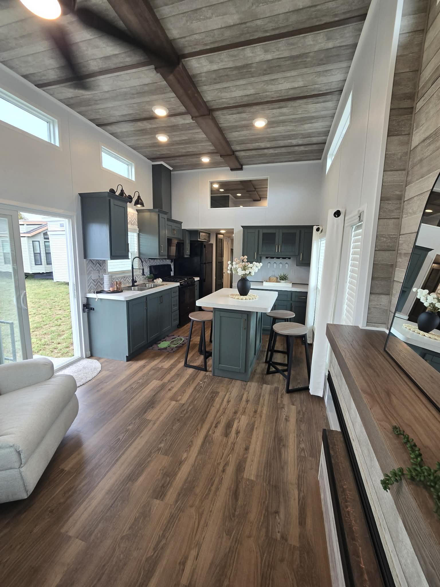 Stylish tiny home interior with wood flooring and a vaulted ceiling. Features a modern kitchen with teal cabinets, island seating, neutral decor, and ample natural light.