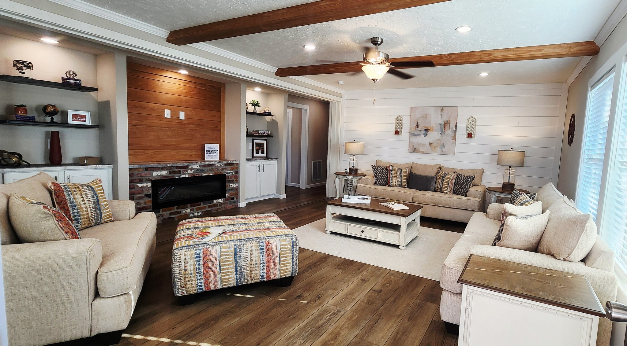 A cozy living room features beige sofas, a striped patterned ottoman, and a wooden coffee table on a light rug. Wooden beams and a ceiling fan enhance the warm, inviting atmosphere.
