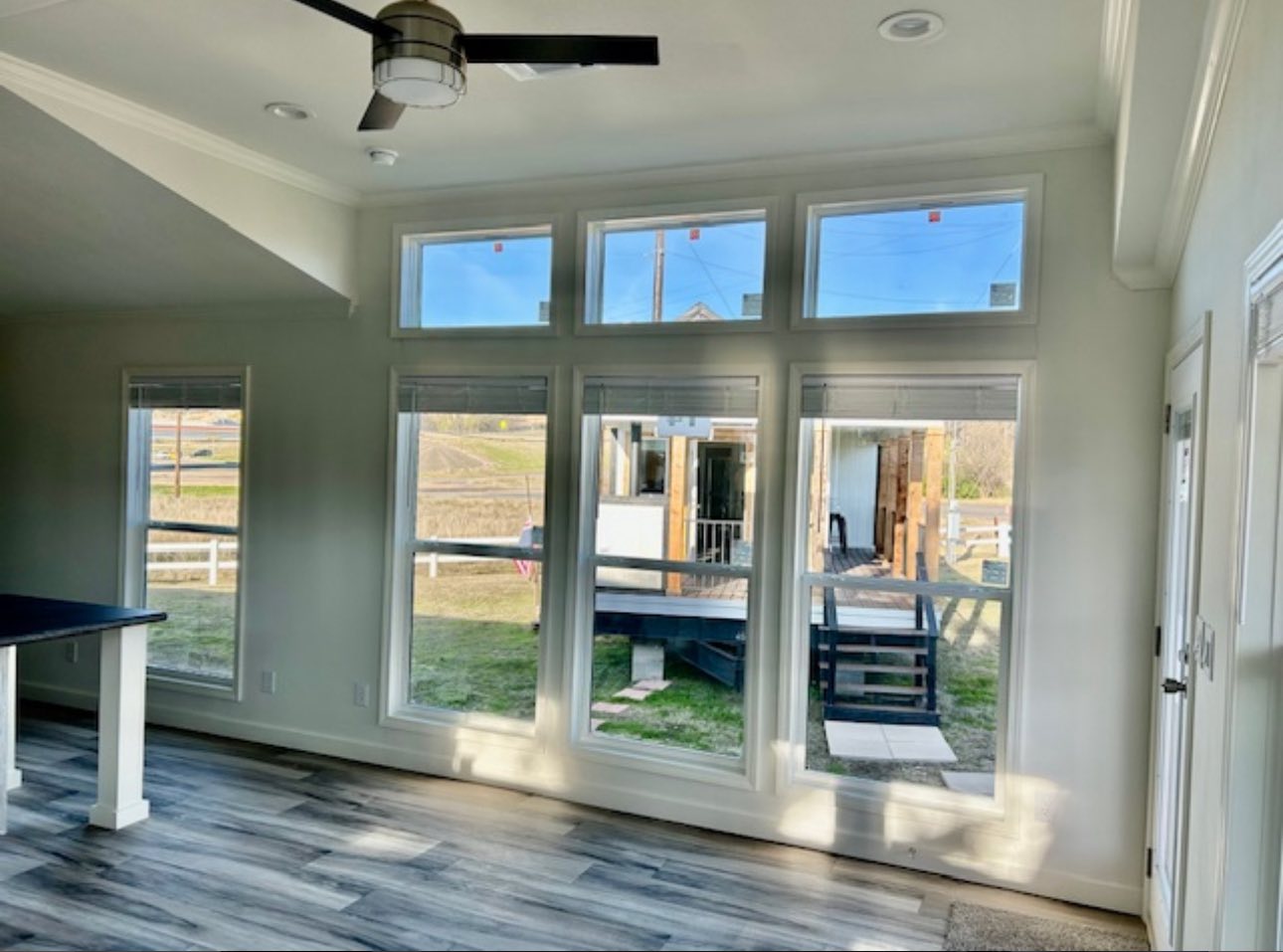 Bright living room with large windows, light streaming in. White walls, wood-like flooring, and a modern ceiling fan create an airy, open feel.