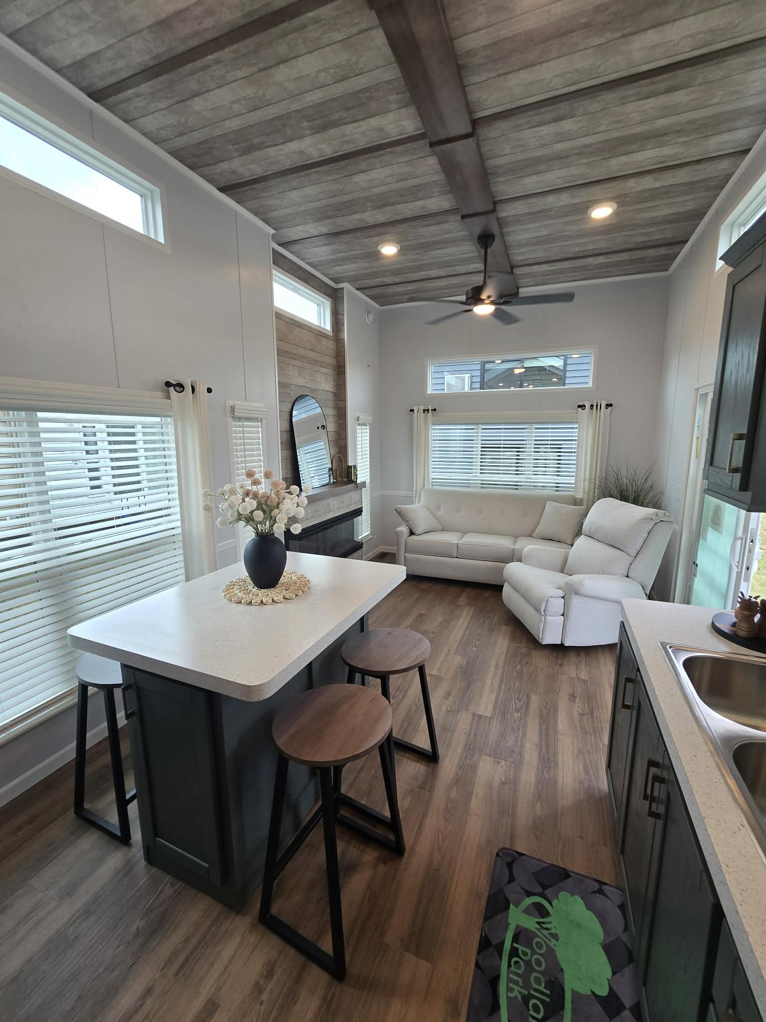 A modern, cozy living area with a high ceiling, wooden floors, a white sectional sofa, island counter with stools, and large windows letting in natural light.