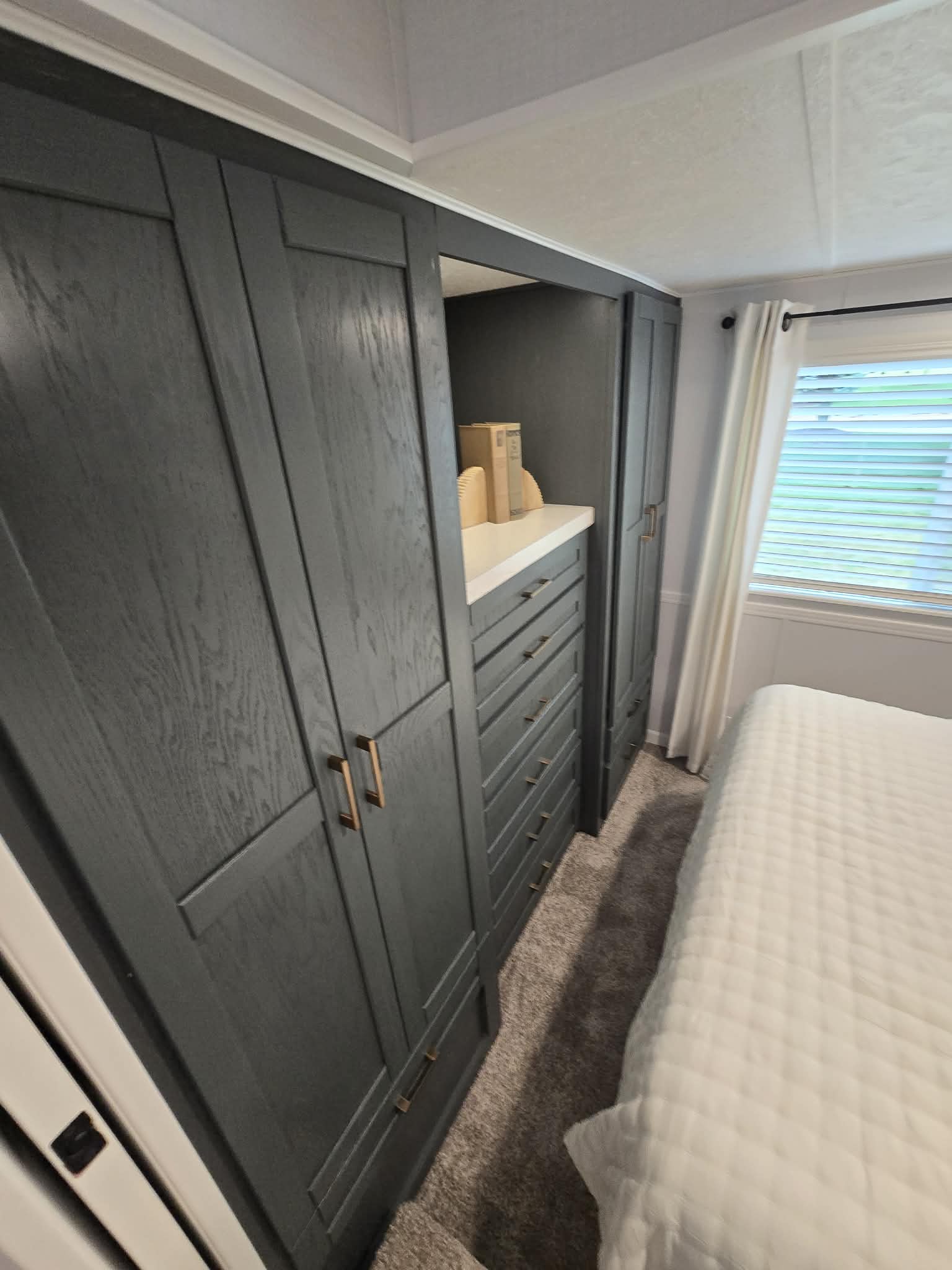 Dark wood wardrobe with brass handles lines a bedroom wall, adjacent to a large window with white curtains. A white bedspread and soft carpet add a cozy touch.
