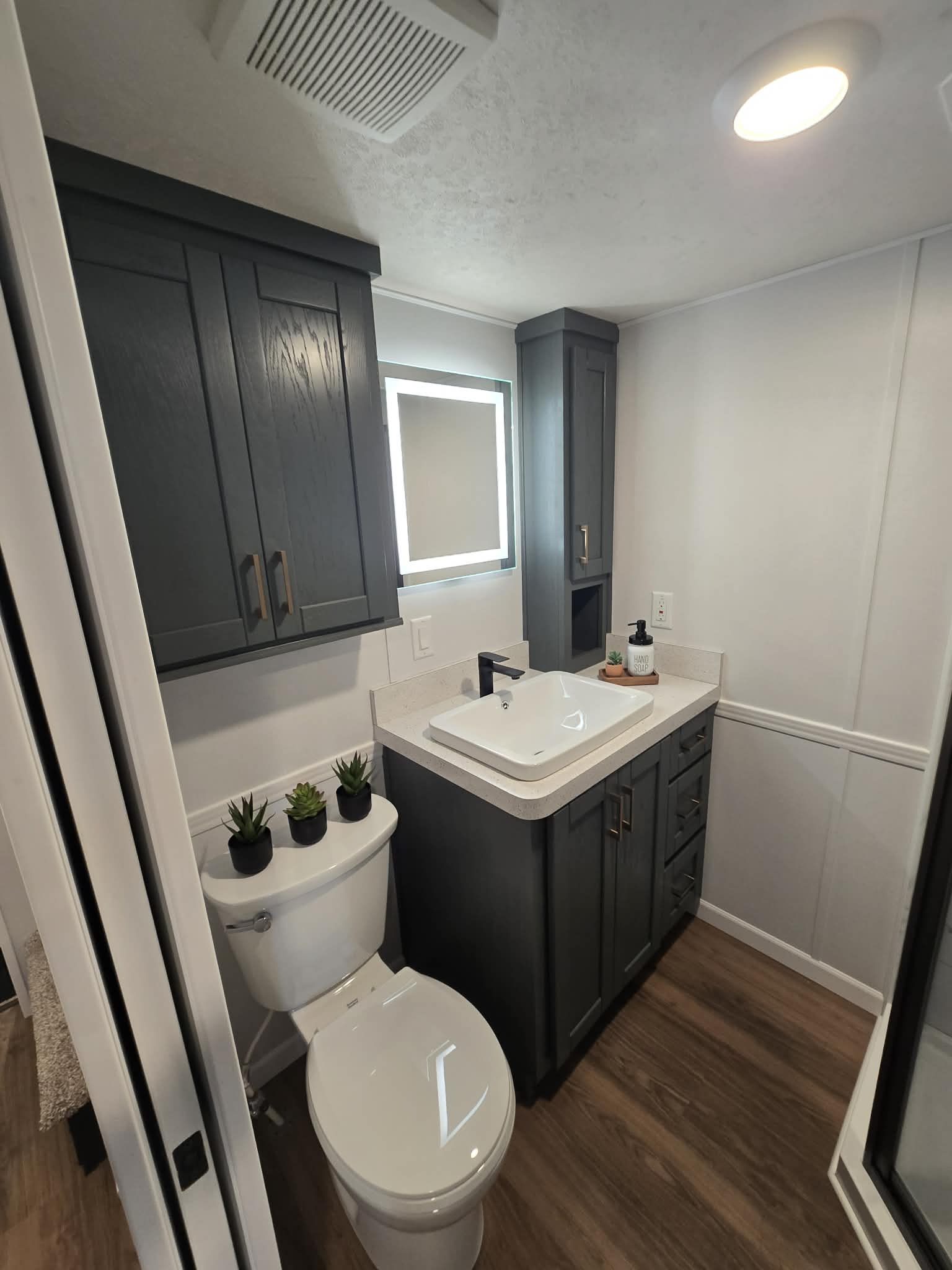 Compact modern bathroom with dark cabinetry and white walls. Features a square sink, wooden floor, small potted plants on the toilet, creating a cozy atmosphere.