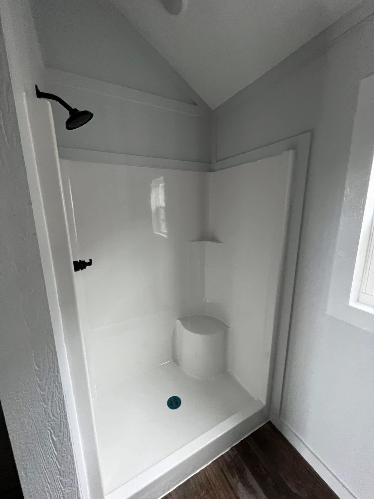 Small, white corner shower with a black showerhead on the left. A simple built-in shelf and seat are attached. Wooden flooring and a bright, minimalist feel.