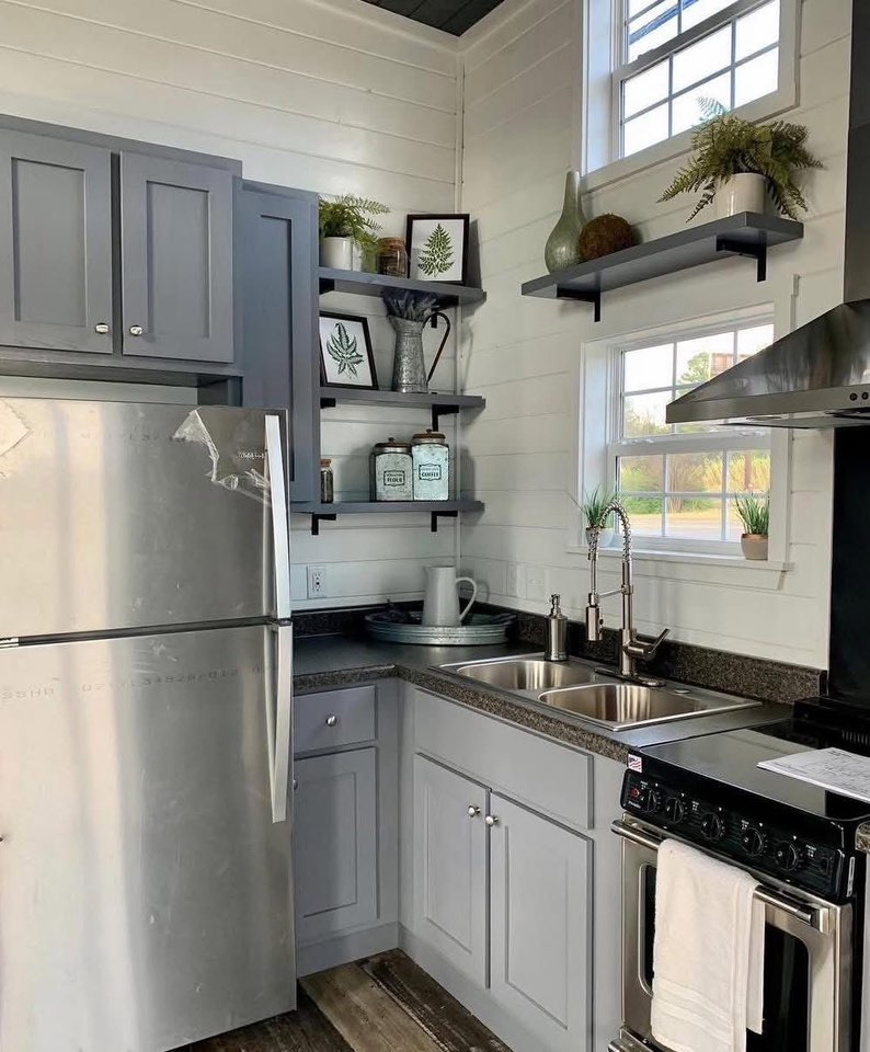 Compact kitchen with gray cabinets, stainless steel fridge, and black countertops. Shelves above hold potted plants and decorative jars, creating a cozy feel.
