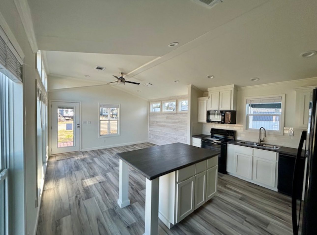 Modern, open-concept kitchen and living area with light wood flooring, white cabinets, black appliances, and a central island. Large windows add brightness.