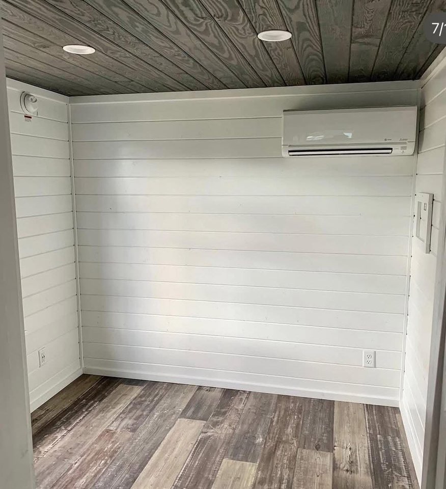 A small room with white shiplap walls, a wooden ceiling with recessed lights, and a sleek air conditioner. The floor is covered with rustic wood planks.