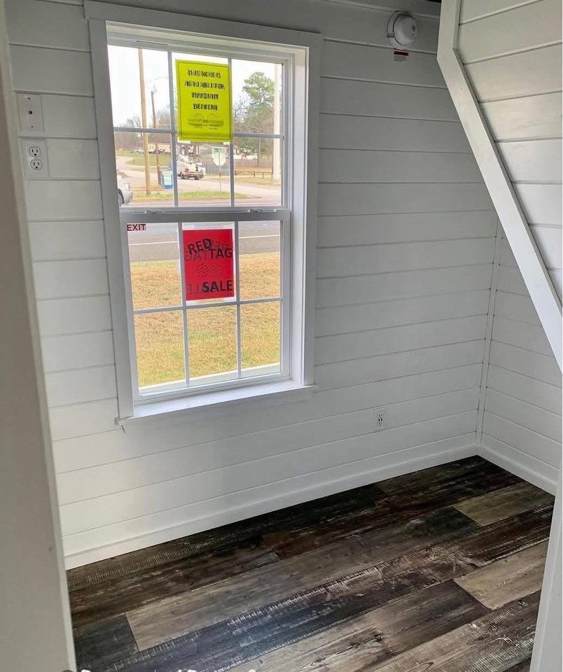 Small white-paneled room with a window displaying red and yellow sale signs. Dark wooden floor adds contrast. View of street and grass outside.