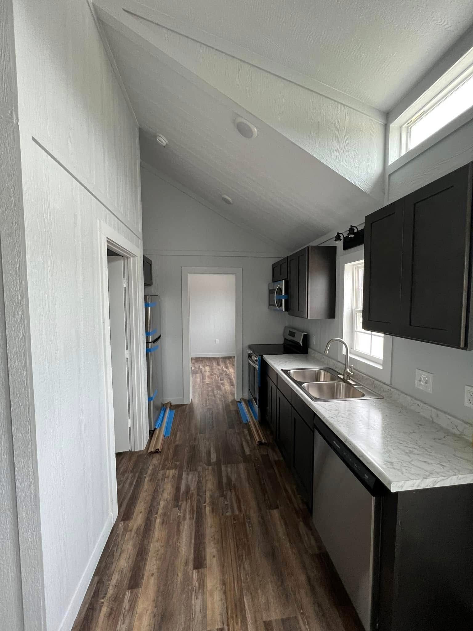 Compact kitchen with vaulted ceiling, dark cabinets, and marble countertop. Wooden floor adds warmth. A bright room is visible through a door in the background.