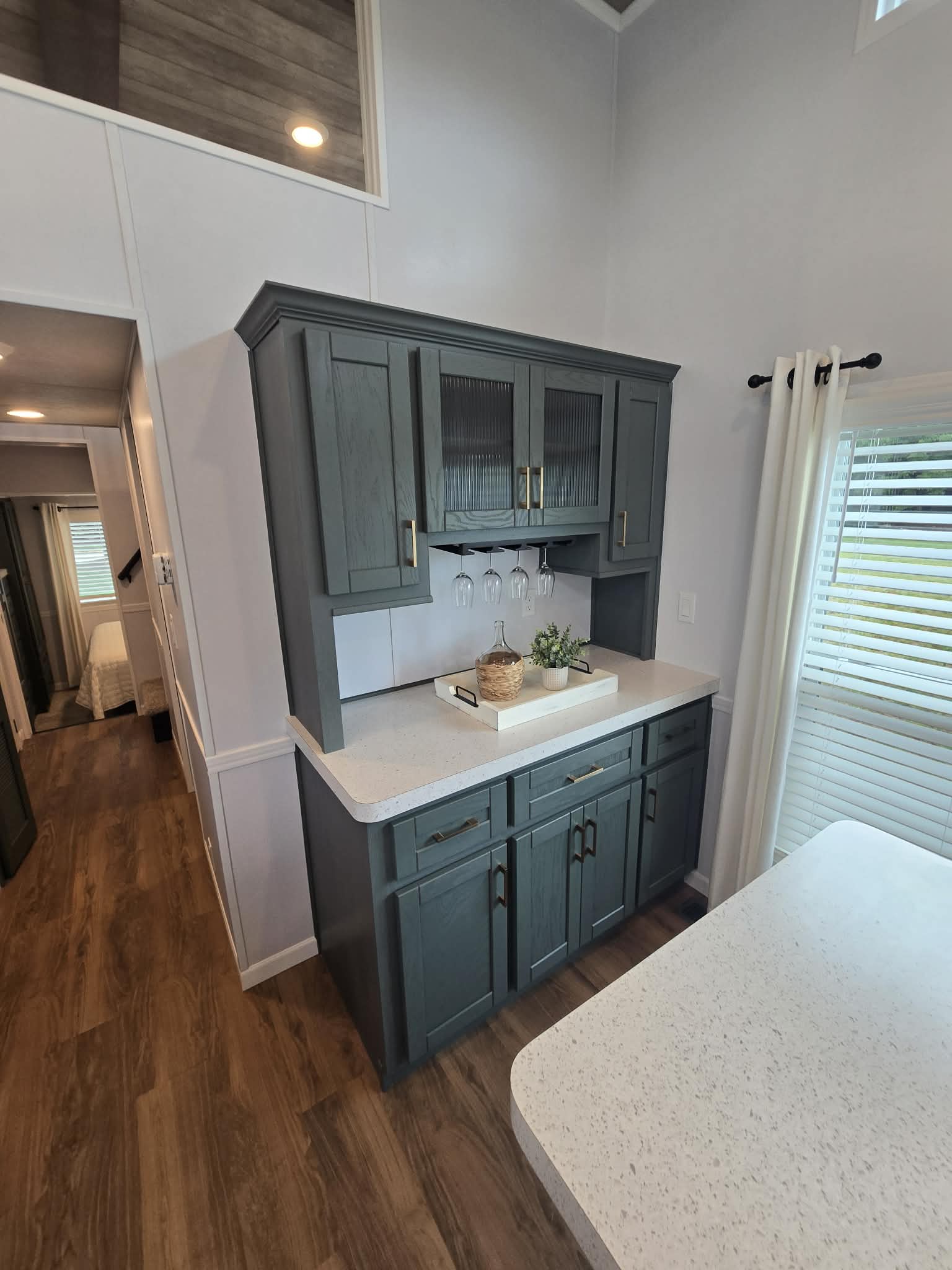 Stylish kitchen nook with dark cabinets, white countertop, and decor tray. Wine glasses hang below the cabinet. Wood flooring adds warmth. Bright, airy feel.