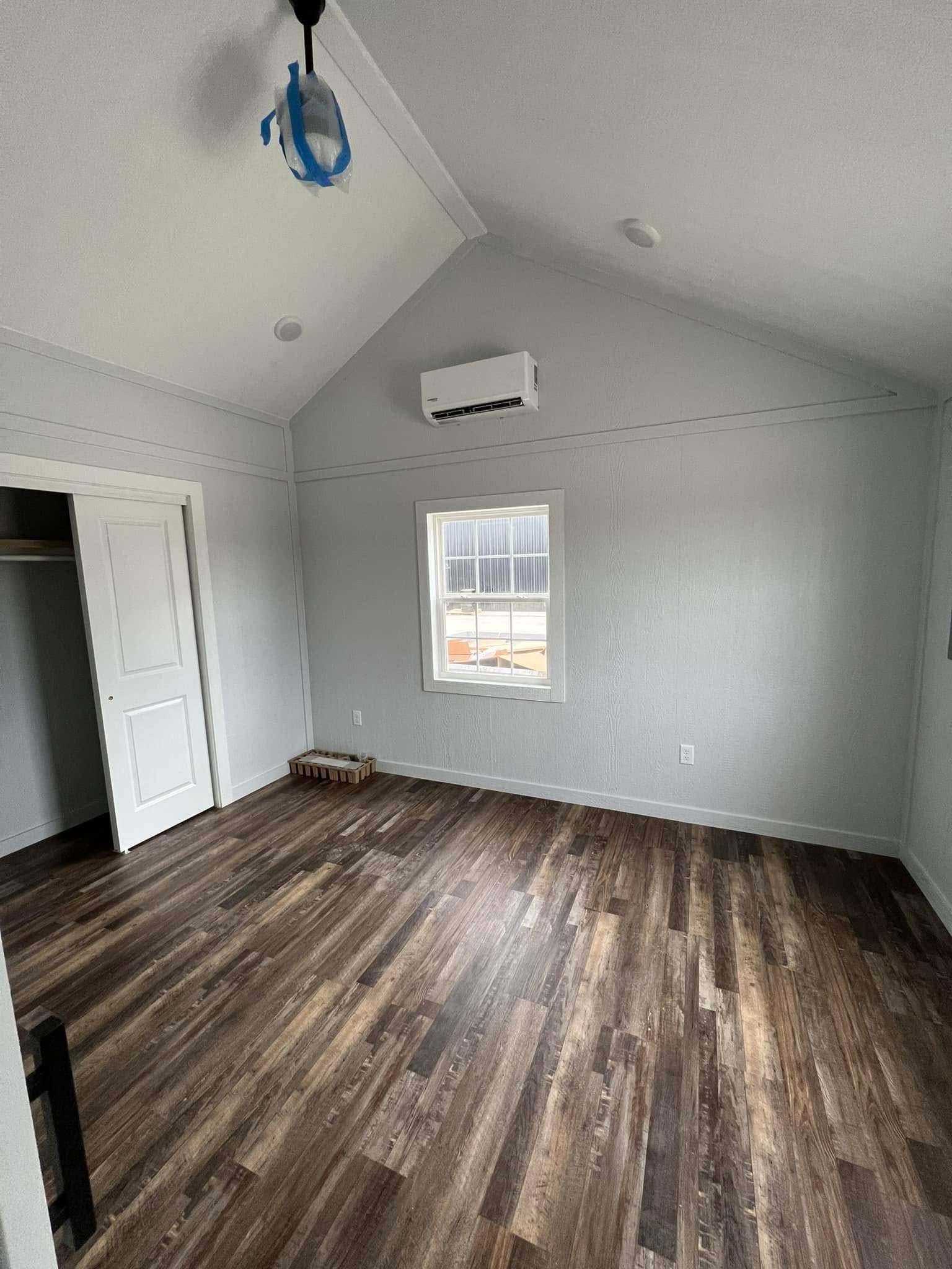 Spacious room with high ceiling, light gray walls, and wood flooring. A window with natural light and an air conditioning unit are present. Minimalist and serene.