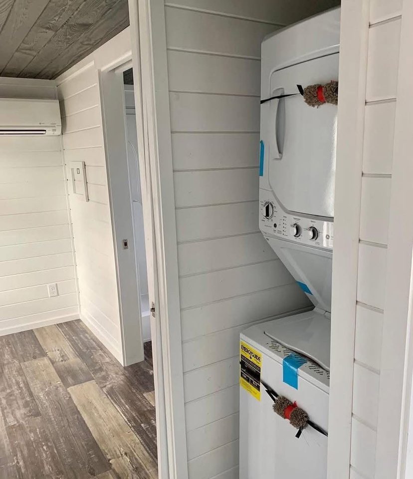 A compact laundry room with a stacked washer and dryer next to a doorway. The walls feature white shiplap, and the floor has a wood-like texture.