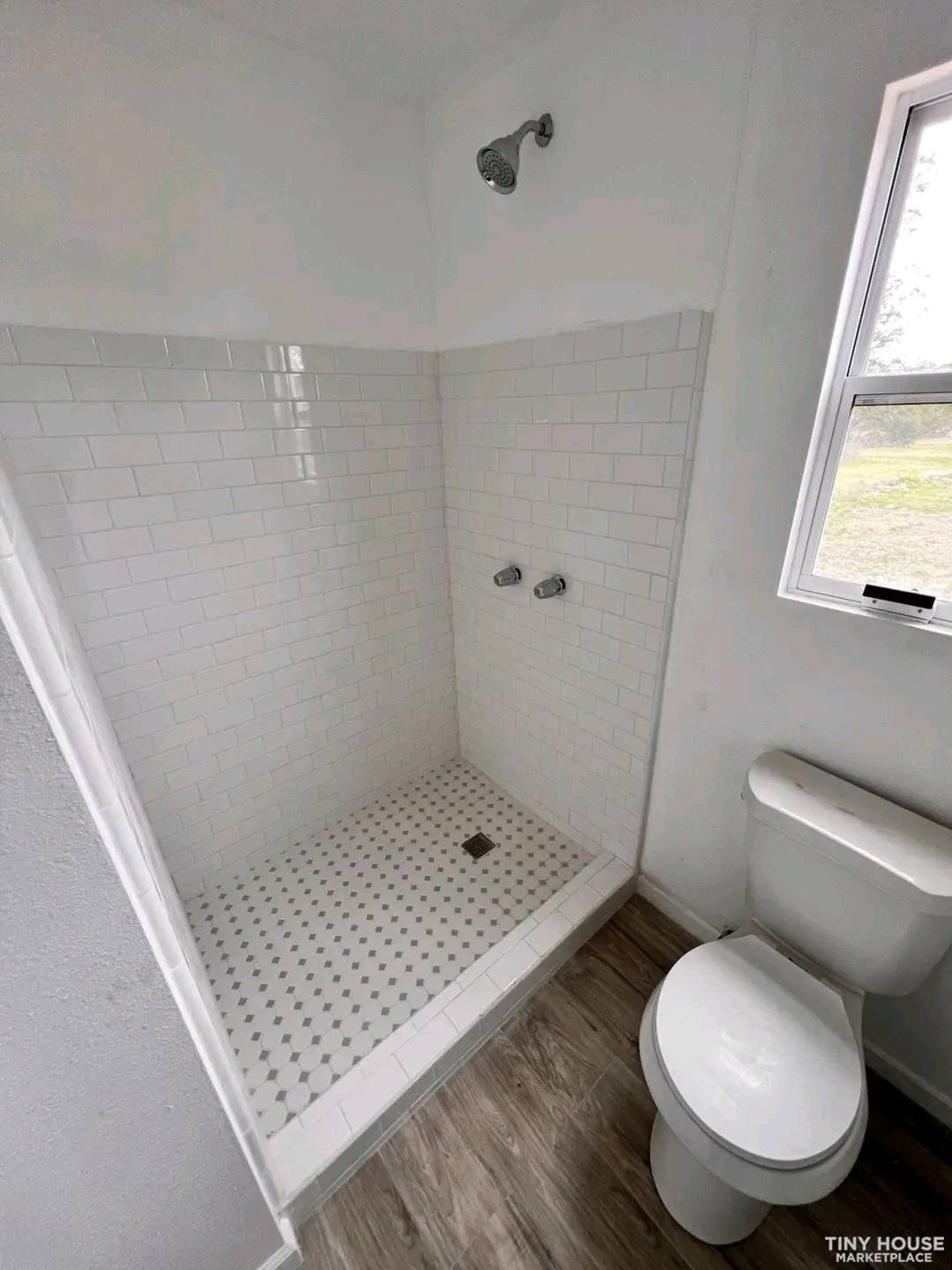 Compact bathroom with white subway-tiled shower, chrome fixtures, and a small window. Adjacent is a toilet on a wood-patterned floor, creating a tidy space.
