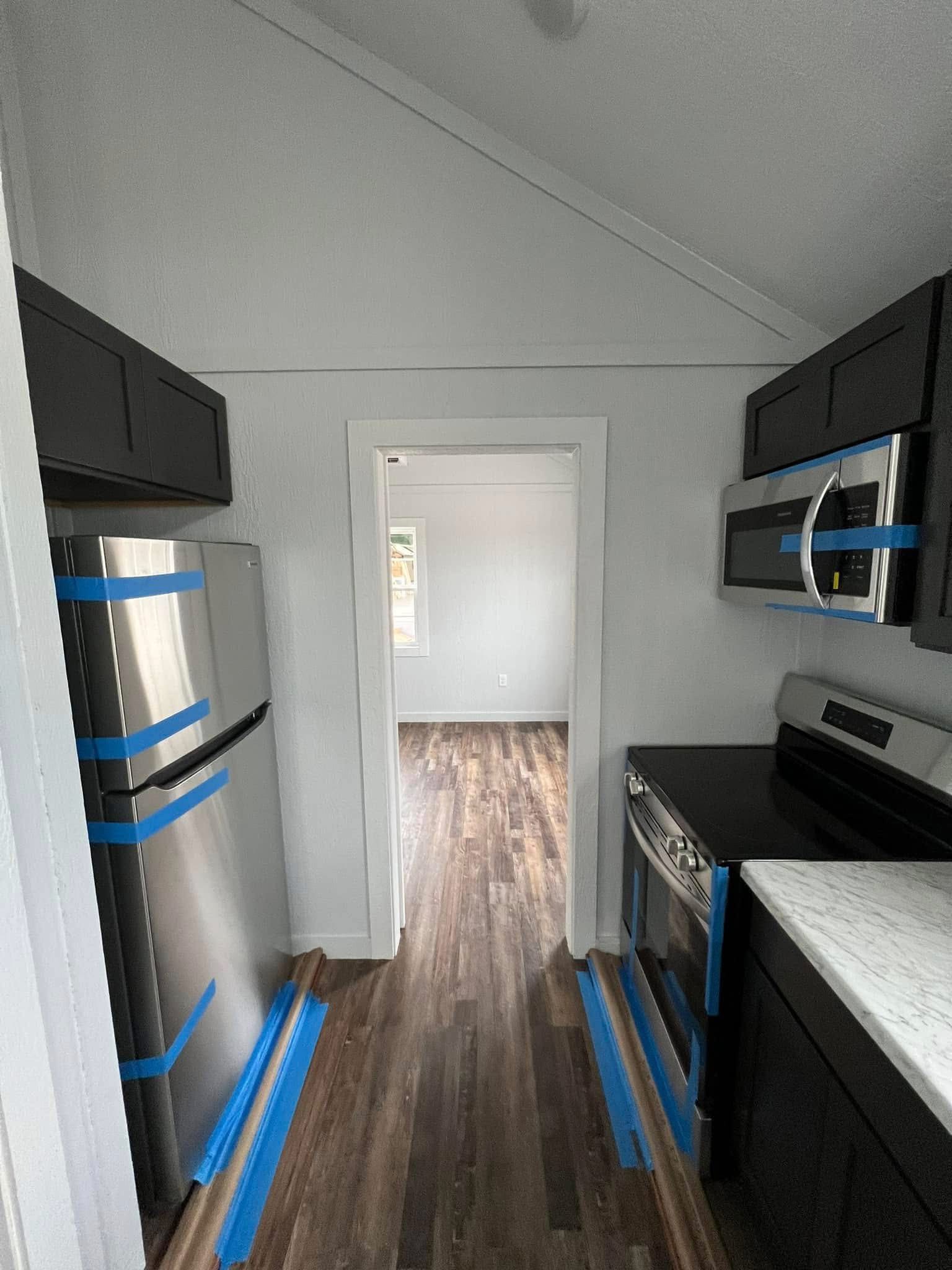 Modern galley kitchen with sloped ceiling, dark cabinets, stainless steel appliances, and blue tape. Wooden floor leads to a bright room. Clean, new.