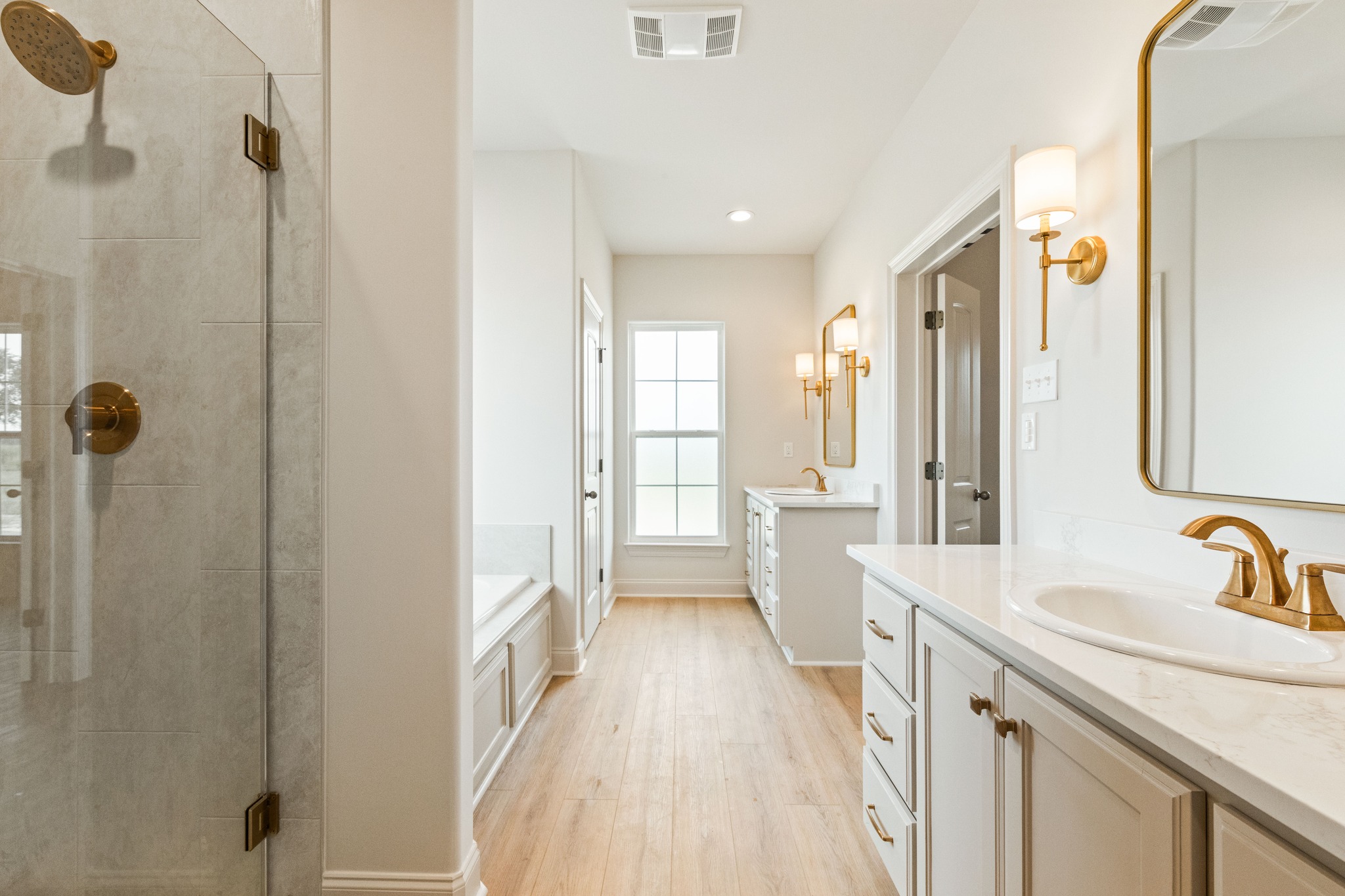 Elegant bathroom with light wood flooring, white cabinets, and brass fixtures. Features include a glass shower, large mirrors, and a window.