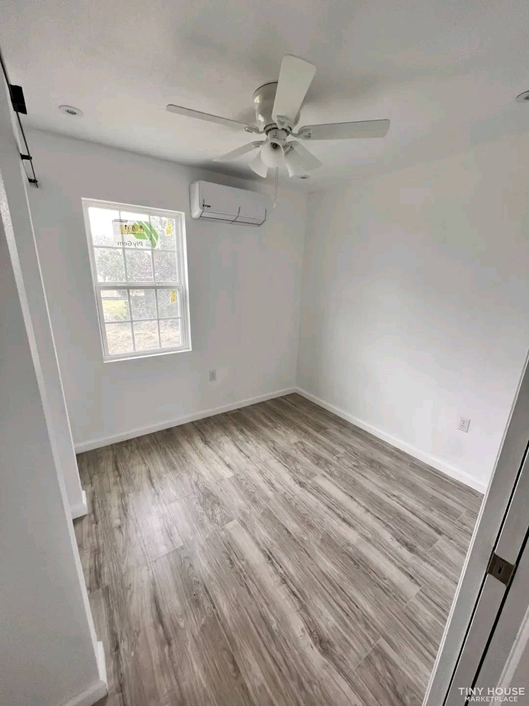 Small, bright room with white walls and light wood floor. Features a window, ceiling fan, and wall-mounted air conditioner, creating a minimalist feel.