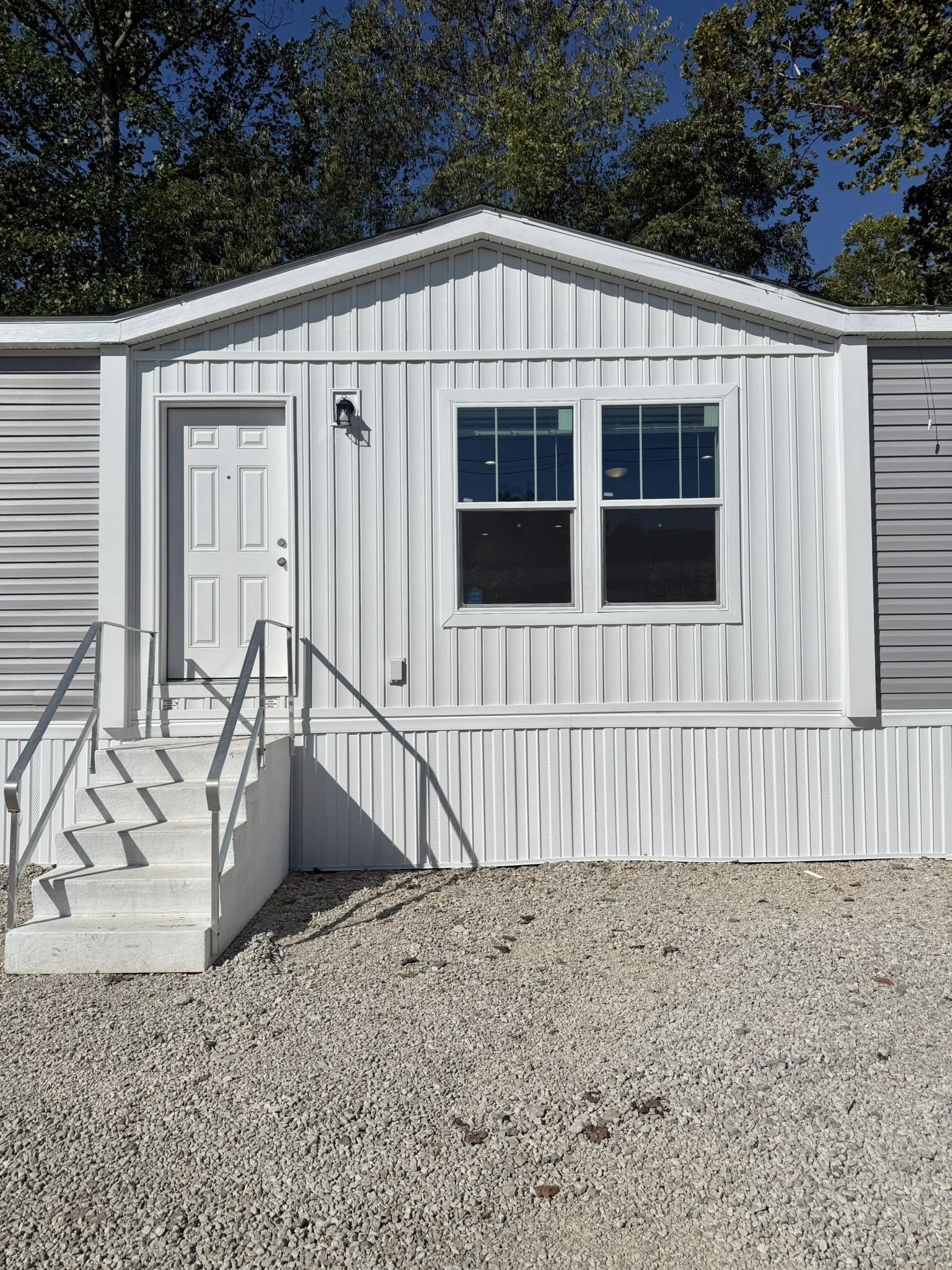 A white mobile home with gray accent, front door, and a pair of large windows. White steps with metal railings lead up to the door. Trees in the background.