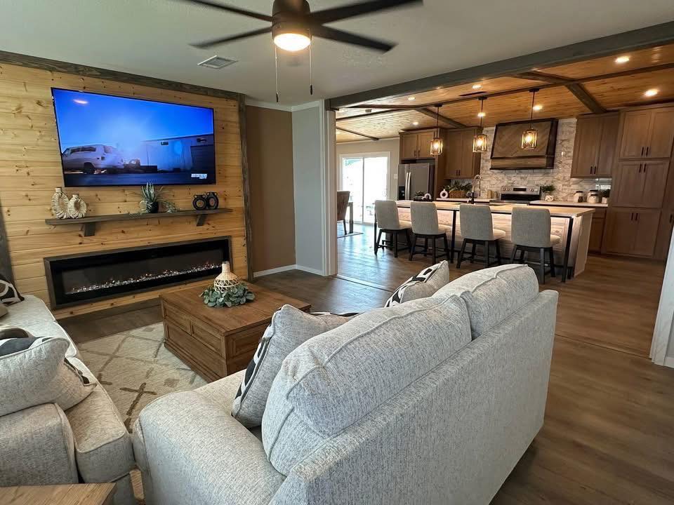 Modern living room with gray sofas and a wood-paneled wall featuring a TV and electric fireplace. Open kitchen with island, stools, and pendant lights. Cozy and inviting.