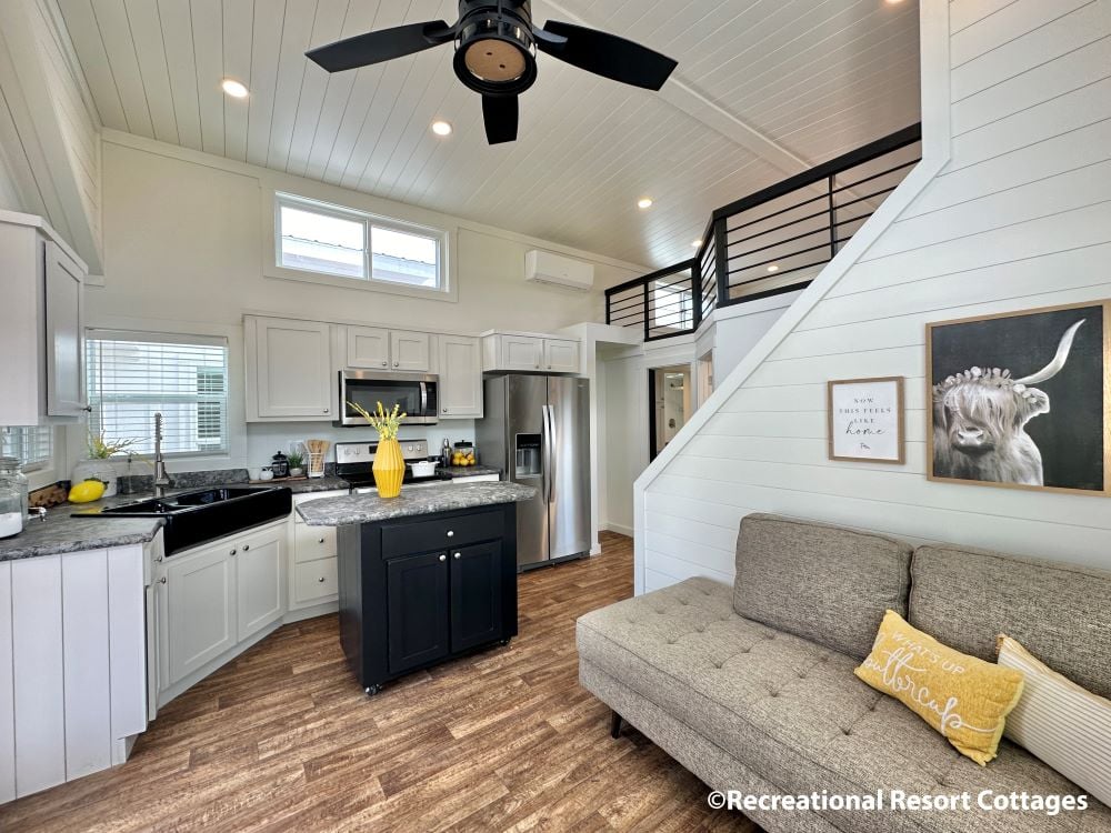 Modern tiny home interior with bright kitchen, featuring white cabinets, black island, and stainless steel appliances. Cozy living area with gray sofa, accent pillows, and wall art. Stairs lead to loft, while wood flooring and ceiling fan complete the airy space.