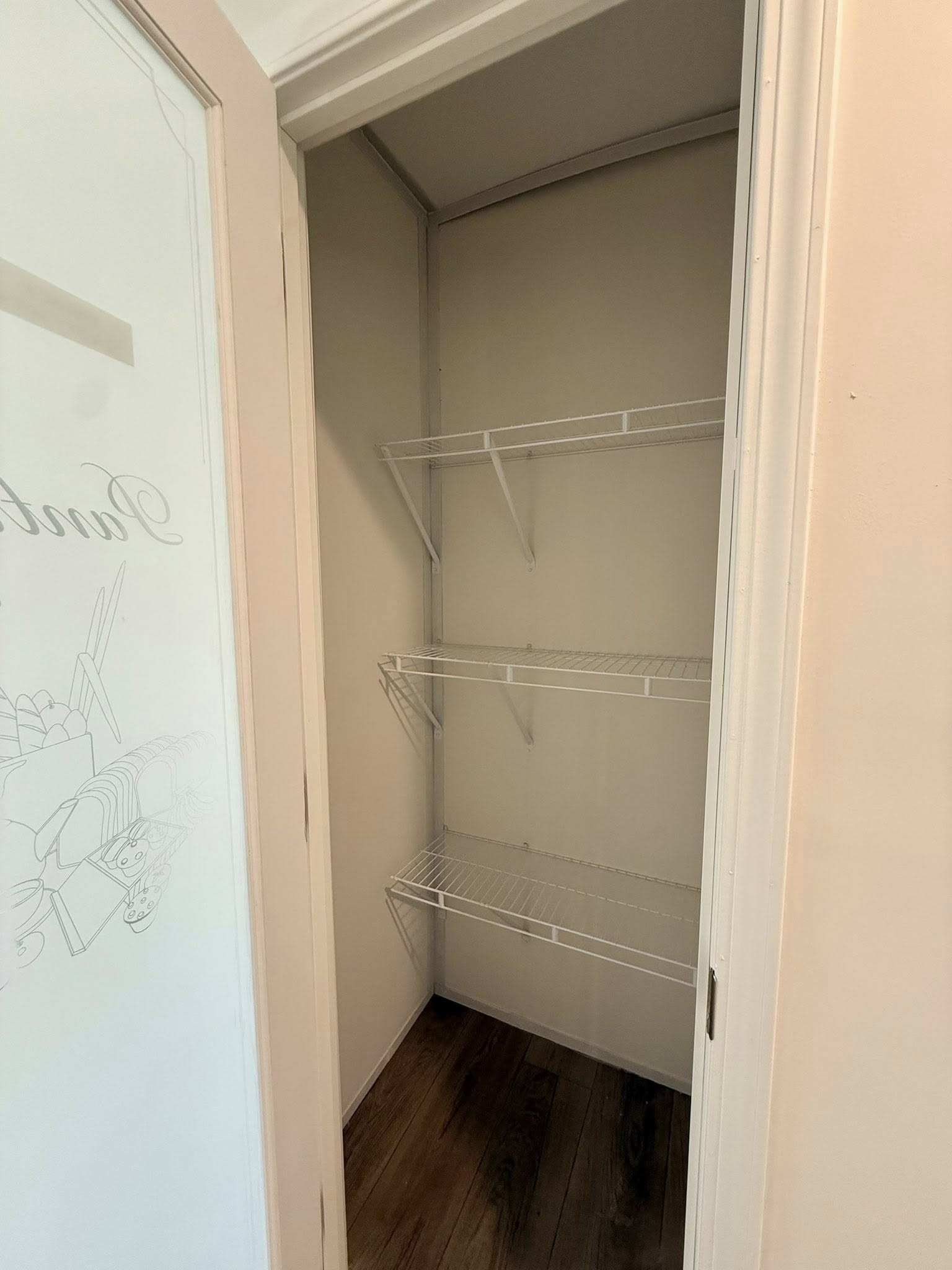 Empty pantry with two white wire shelves, wooden floor, and light beige walls. Glass door has subtle food illustrations and text. Minimalist style.