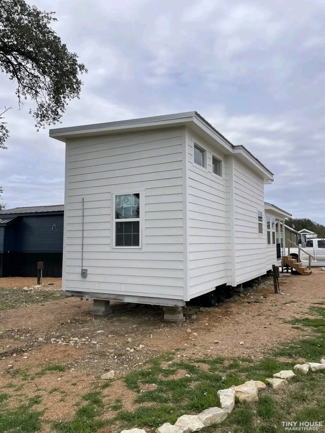A modern, white tiny house with horizontal siding sits elevated on blocks. It has windows and a simple, compact design, surrounded by a gravel path and patches of grass under a cloudy sky.