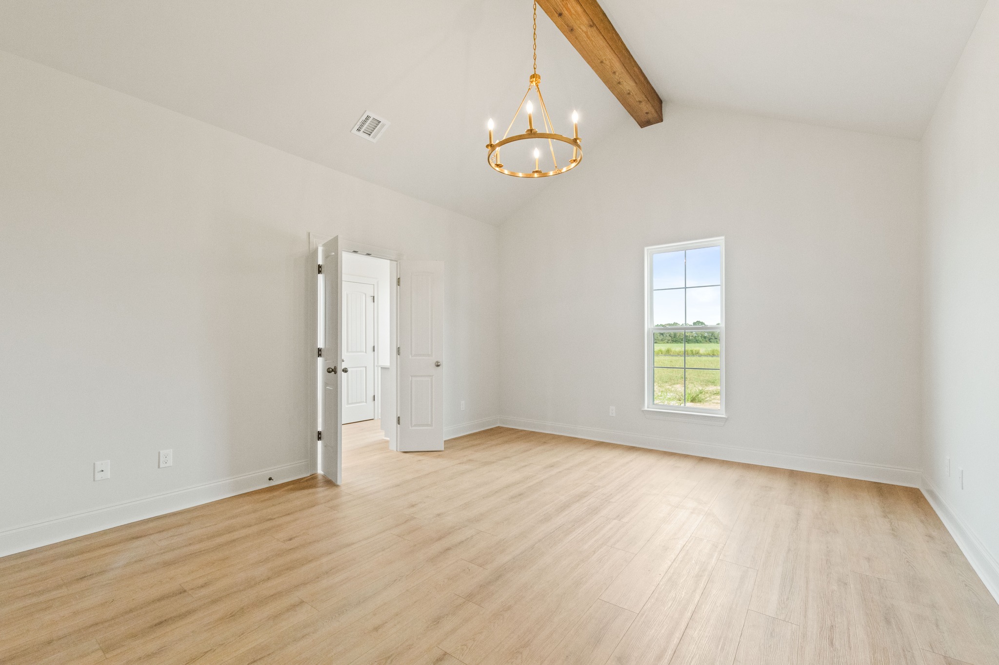 Spacious room with light wood flooring, a vaulted ceiling with wooden beam, large window with scenic view, modern chandelier, and open white doors.