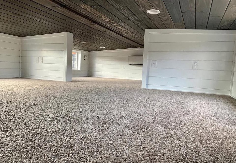 A small, cozy attic space with white paneled walls and a low dark wooden ceiling. The carpeted floor adds warmth, with small windows providing light.