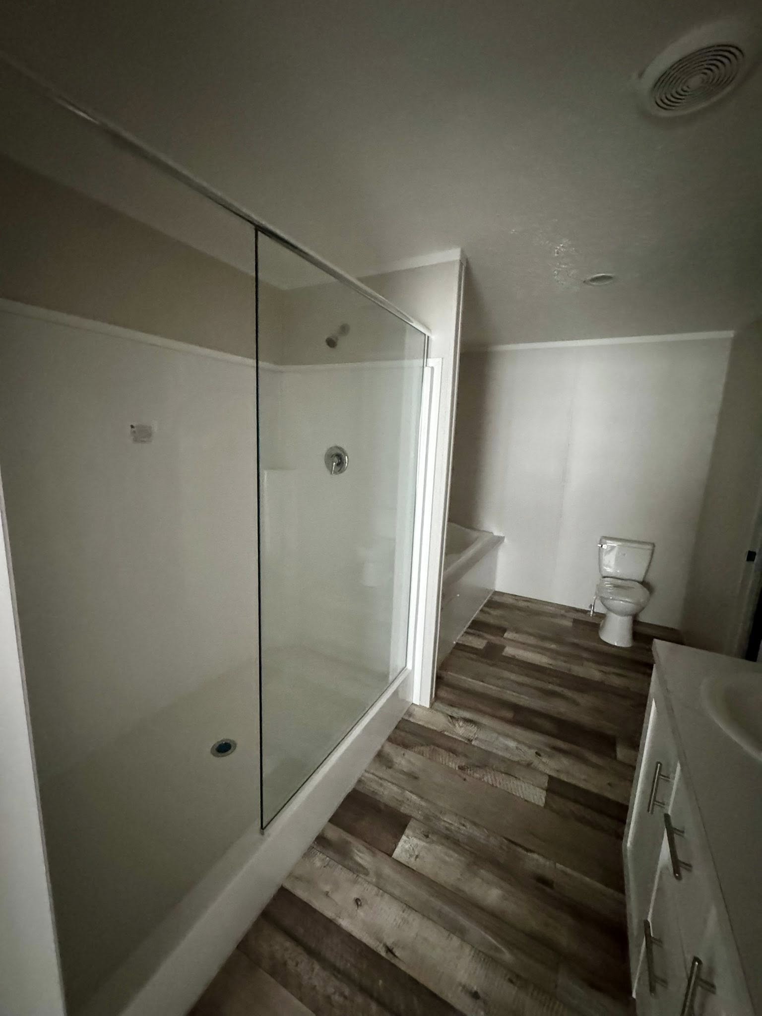 A modern bathroom featuring a glass-enclosed shower on the left, a wooden floor, white cabinets with silver handles, and a toilet in the back right.