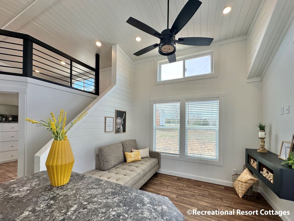 Spacious modern living area with high ceilings, featuring a gray sofa, yellow vase with flowers, ceiling fan, large windows, and decorative items. Cozy and inviting.