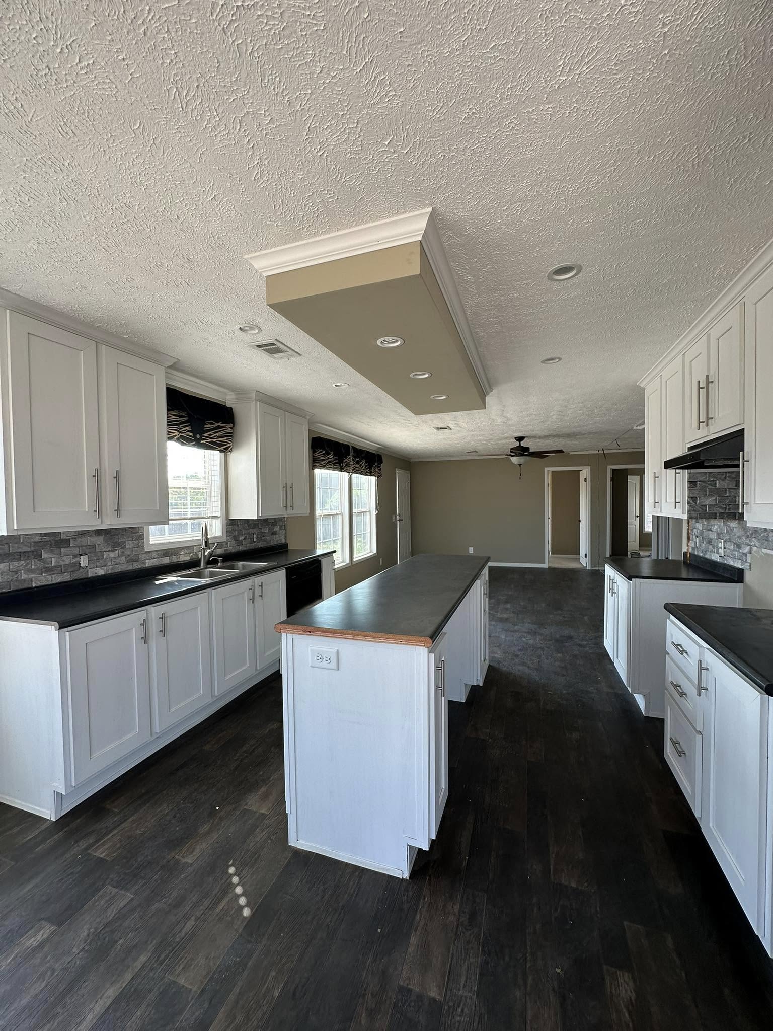 Spacious kitchen with dark wood floors, white cabinets, and black countertops. Central island, ceiling fan, and large windows. Bright, modern ambiance.