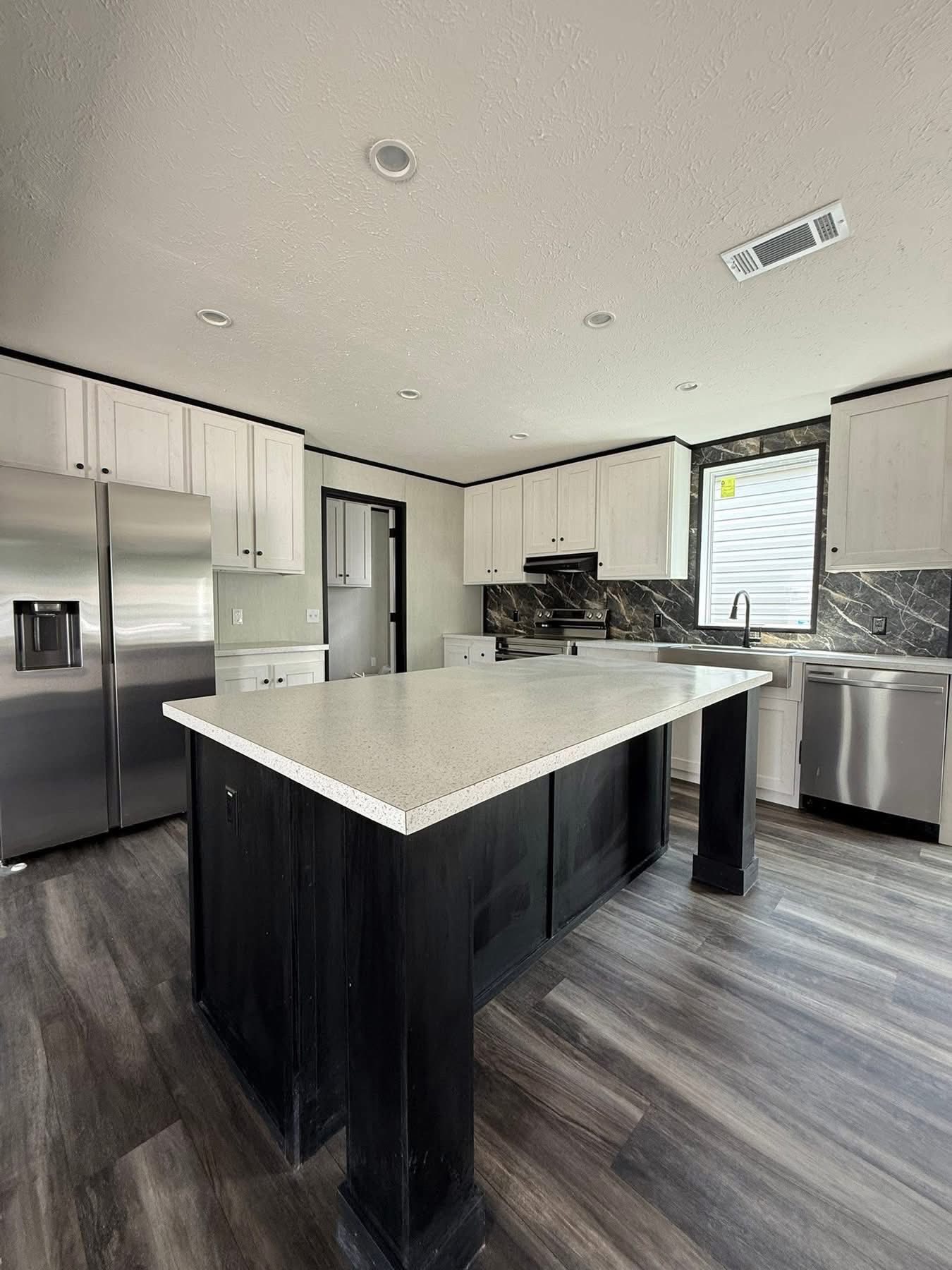 Modern kitchen with large island, light countertops, dark wood base, stainless steel appliances, and white cabinets. Warm lighting and sleek flooring create a clean and inviting atmosphere.