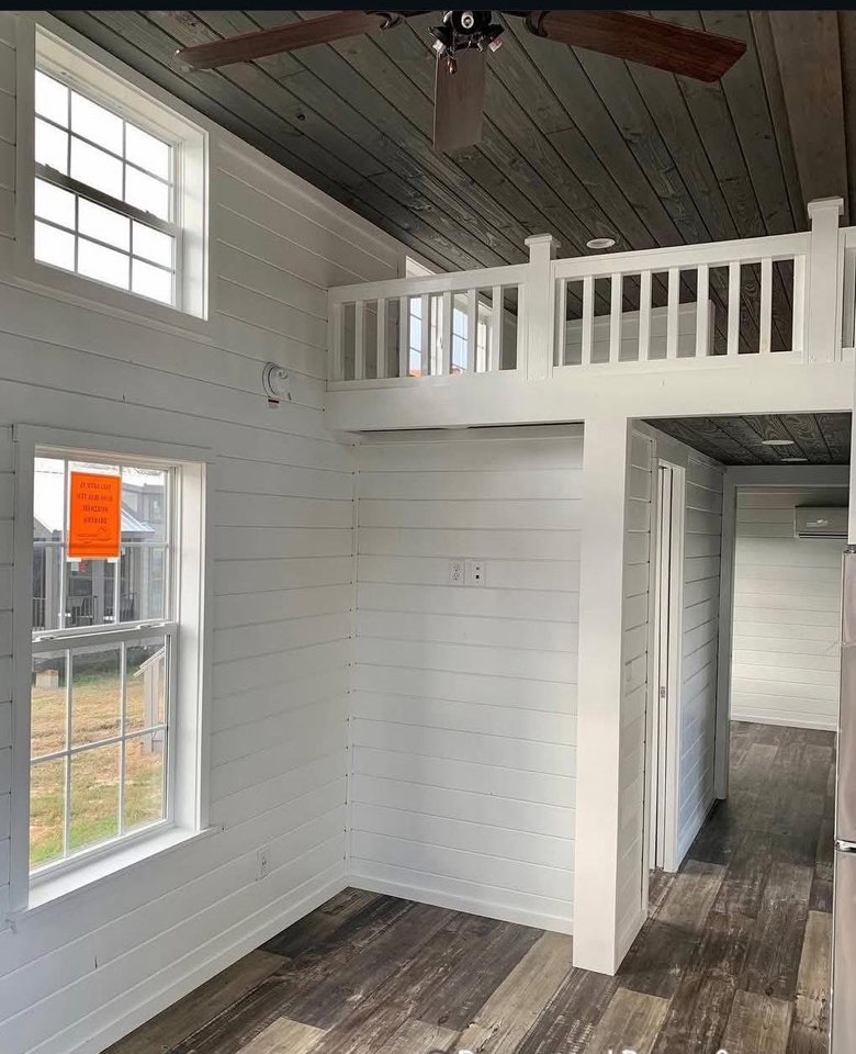 Small room with white wood panel walls, a dark wood ceiling, and brown vinyl flooring. Natural light enters through windows; a loft space is visible above.
