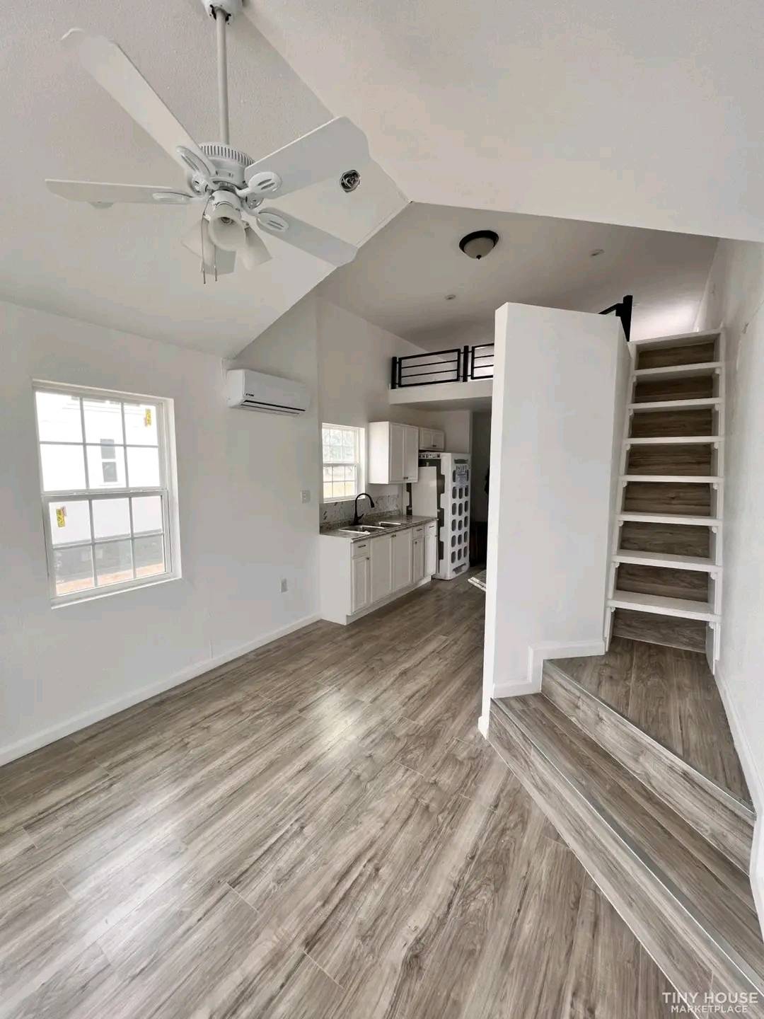Bright tiny house interior with light wood flooring, white walls, and ceiling fan. Kitchen area with white cabinets and small loft above stairs.