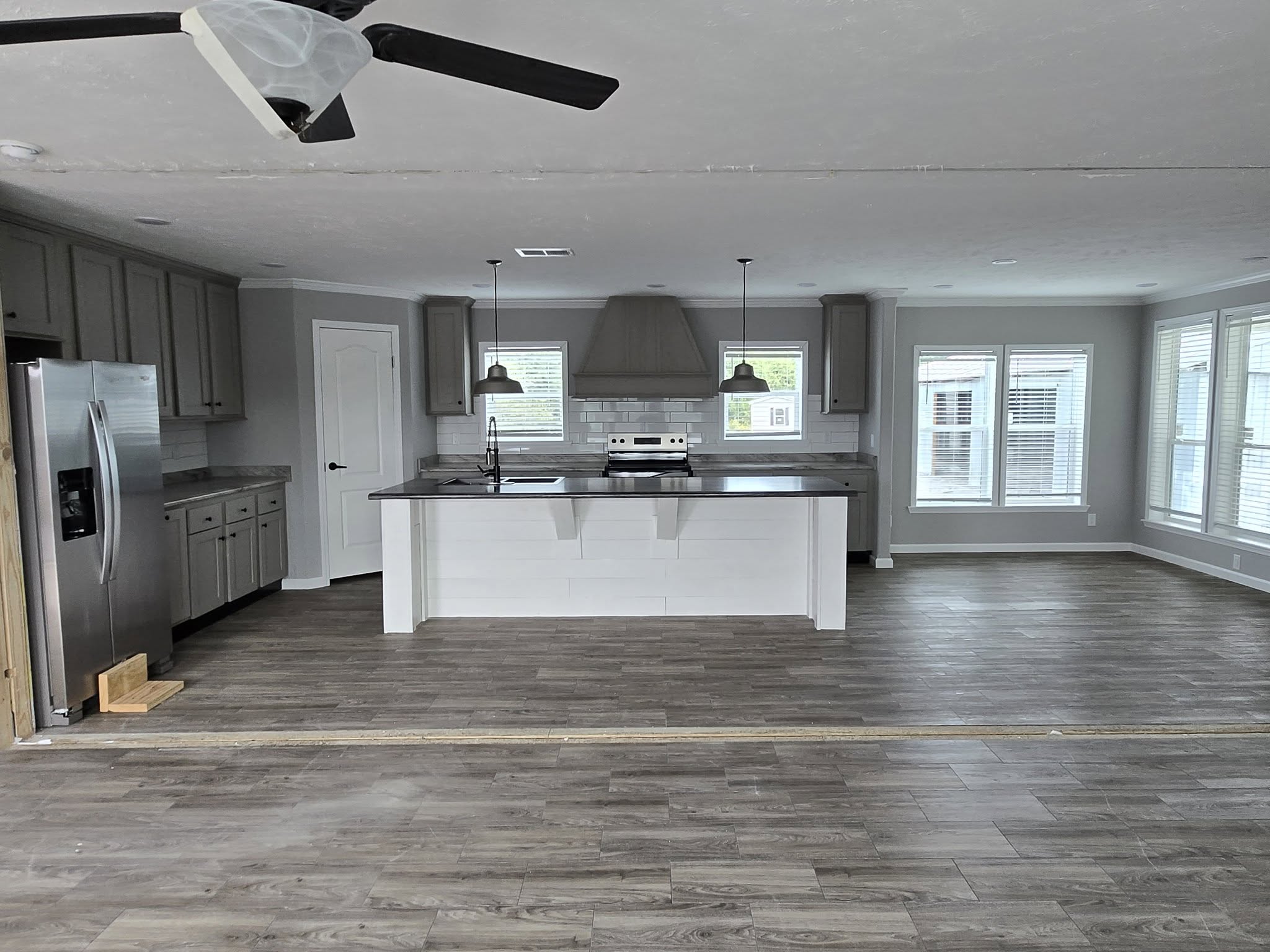 Spacious modern kitchen with gray cabinets, stainless steel fridge, large island, pendant lights, and wood flooring. Bright windows enhance the airy feel.