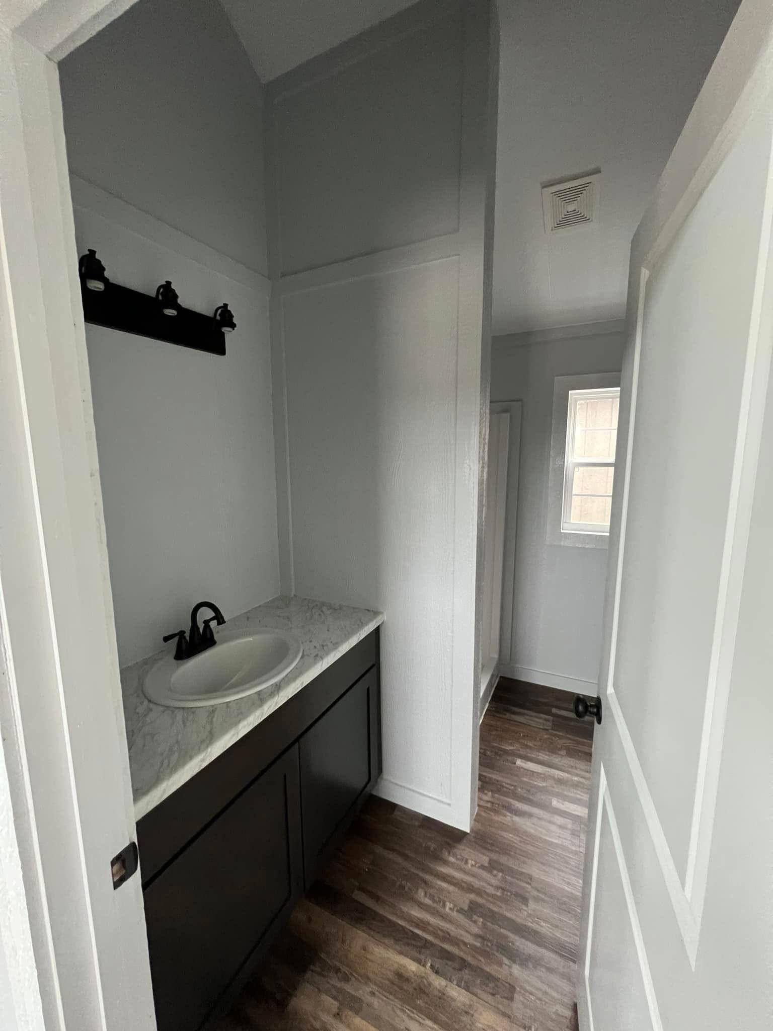 Narrow white bathroom with dark wooden vanity, marble countertop, and black faucet. Single window on the right, wood-textured floor. Modern and minimalistic.