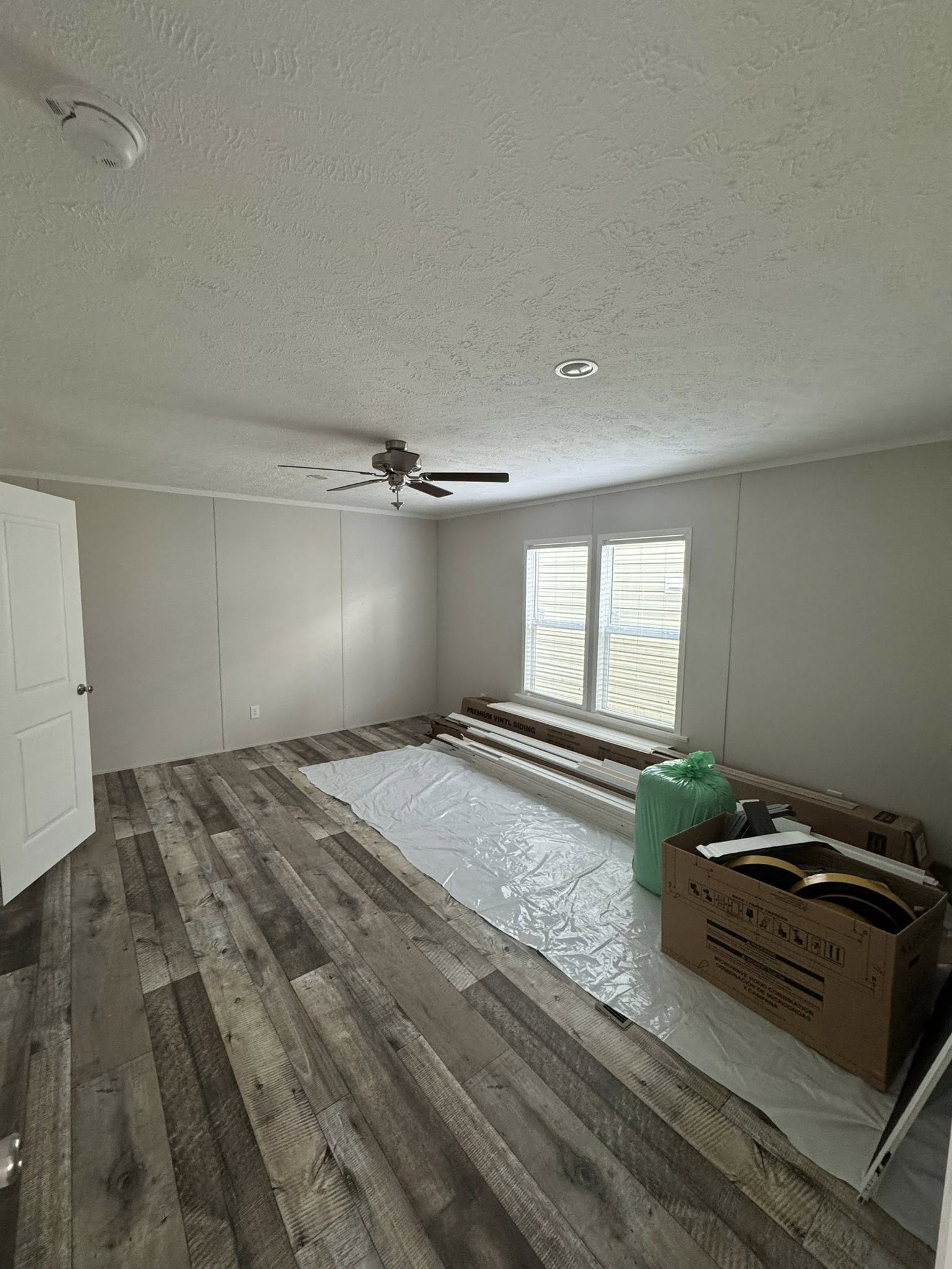 Unfurnished room with wood-patterned flooring, a ceiling fan, and two bright windows. Packing materials and a box are on the floor, indicating a move.