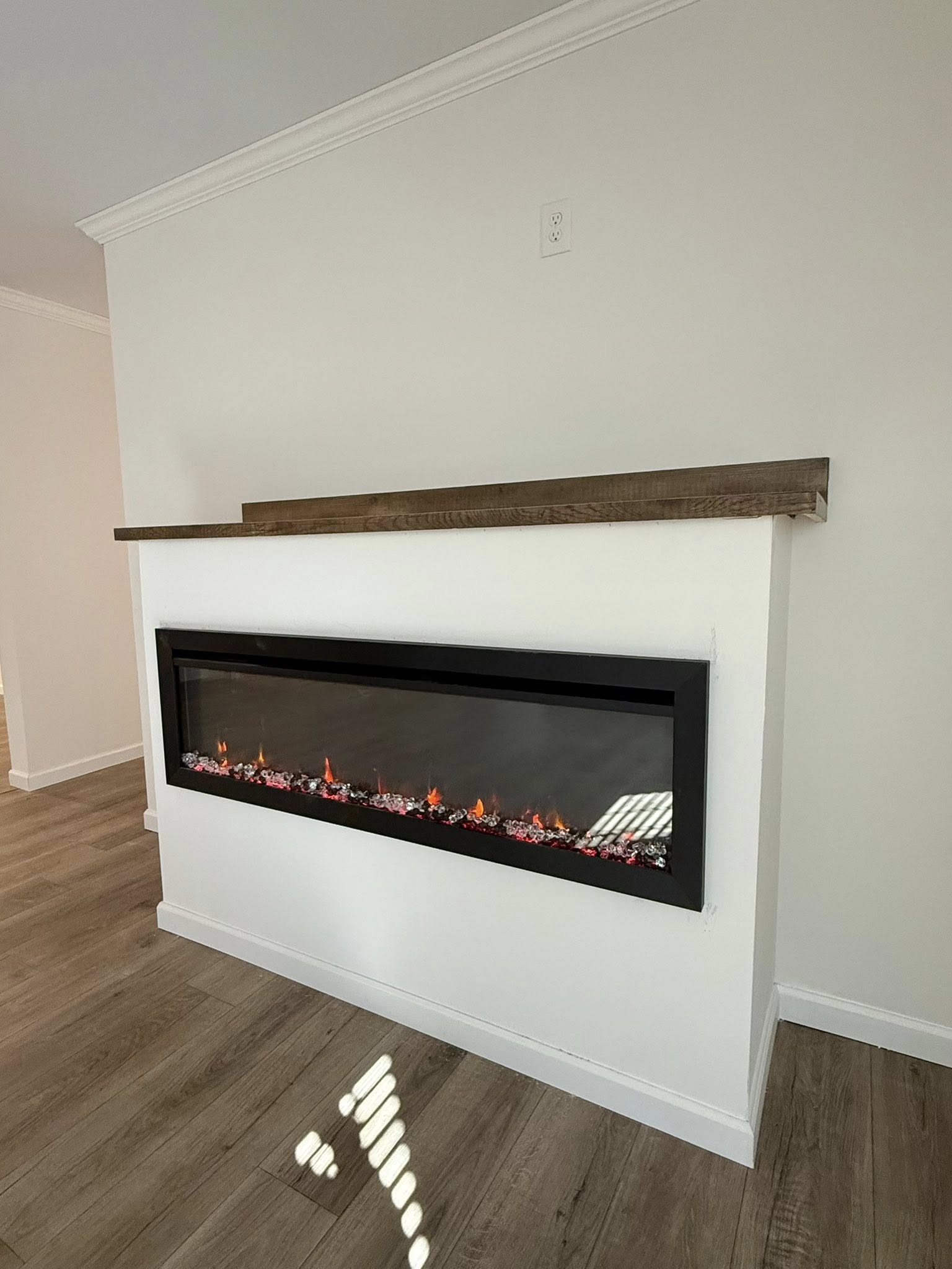 Modern electric fireplace with glowing embers is set in a sleek white wall. A rustic wooden shelf sits above. The atmosphere is cozy and contemporary.