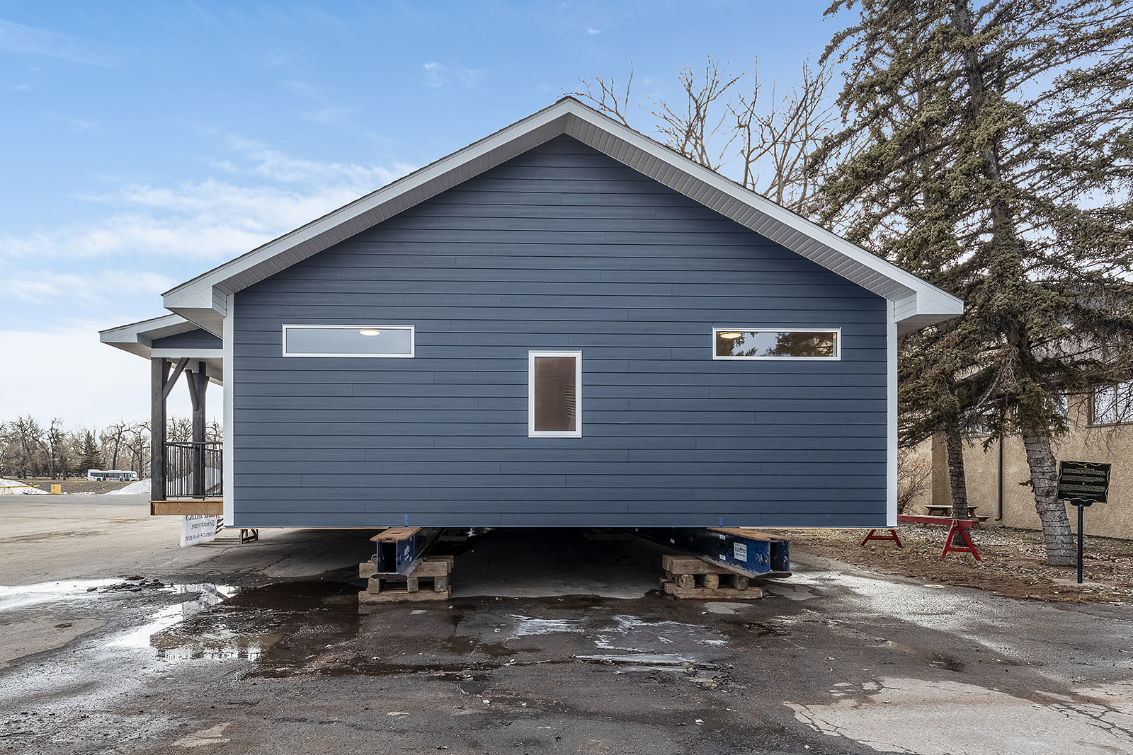 Front view of a blue modular home elevated on pallets, against a cloudy sky. Surrounded by bare trees, the scene feels quiet and industrial.