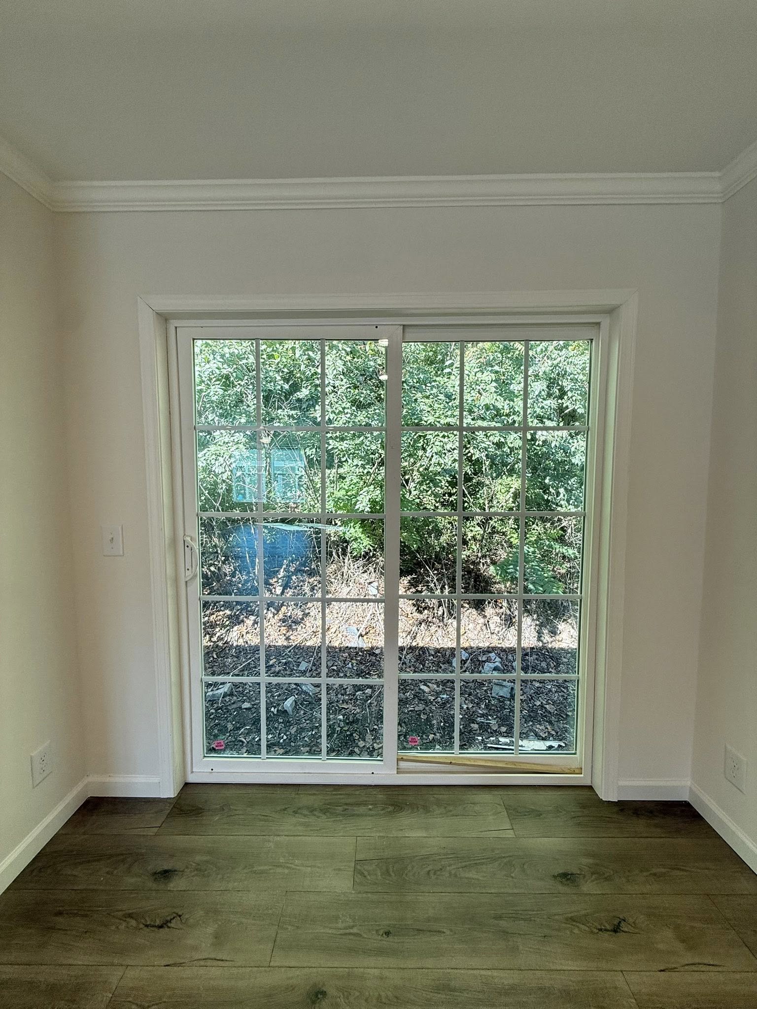 Sliding glass door with white frame, leads to outdoor greenery. Wooden floor inside, light beige walls enhance the room's bright, airy feel.