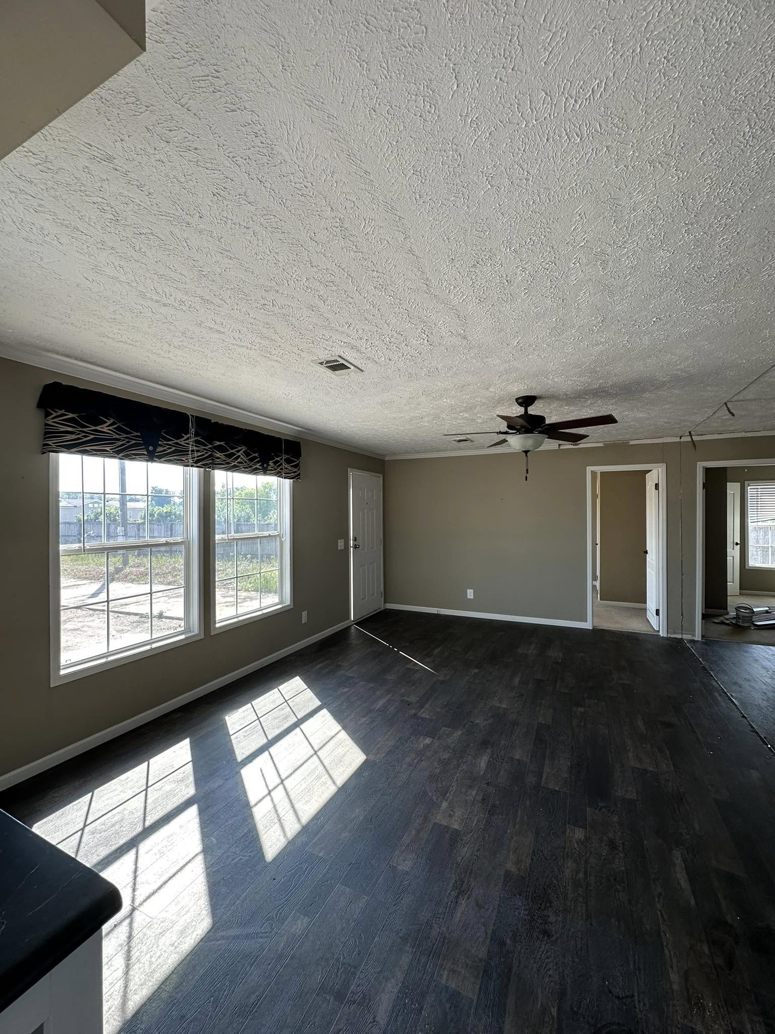 Sunlit empty room with dark wood flooring, beige walls, and a large window casting shadows. Ceiling fan, open doors, bright, airy atmosphere.