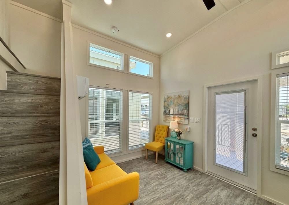 Bright, modern living room with a high ceiling. Features a yellow sofa, teal side table, yellow chair, stairs, and artwork; natural light floods in.