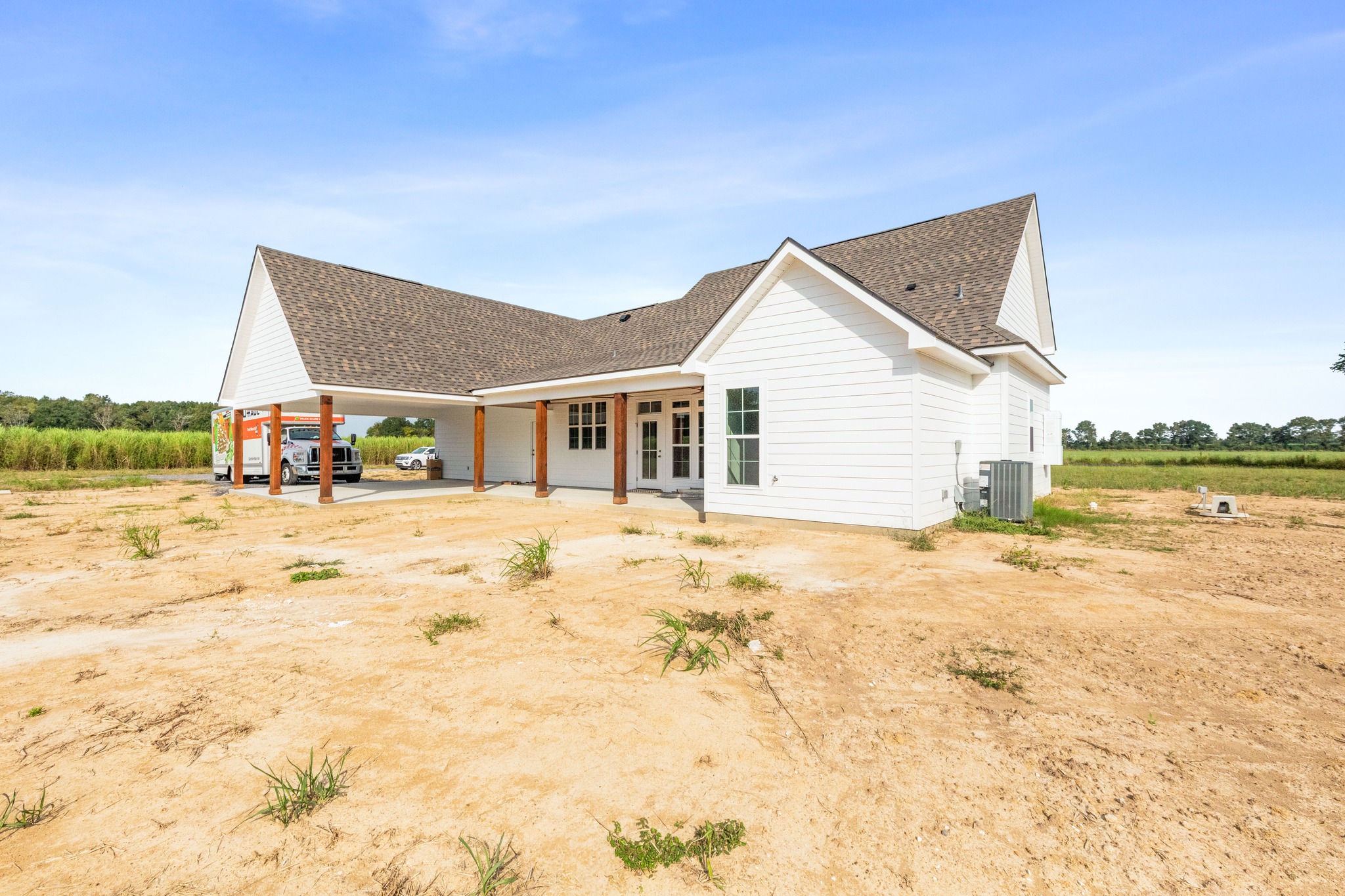 Single-story white house with a wooden porch on a sandy, open plot under a clear blue sky. A parked car is visible under a covered section.