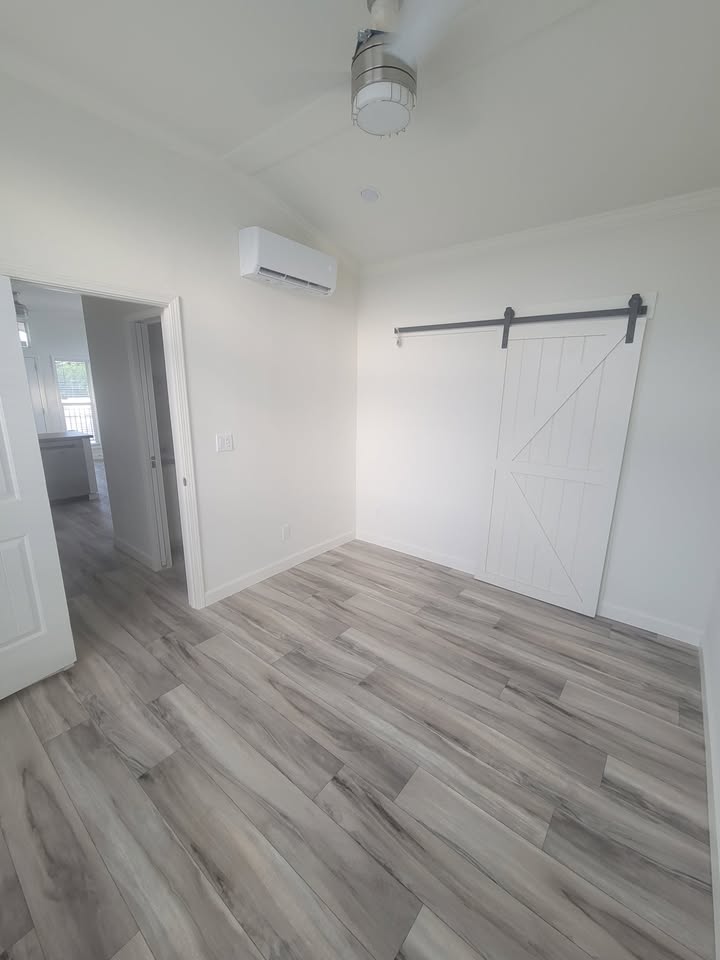 Empty room with light wood flooring, white walls, and a barn-style sliding door. Ceiling fan and air conditioner add modern touches, conveying simplicity.
